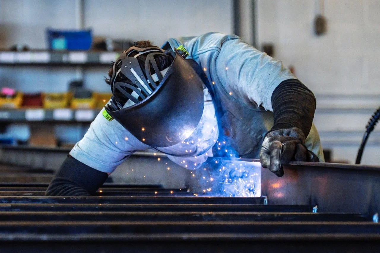 A person wearing a welding helmet, gloves, and protective clothing welds metal, creating sparks in an industrial workshop.