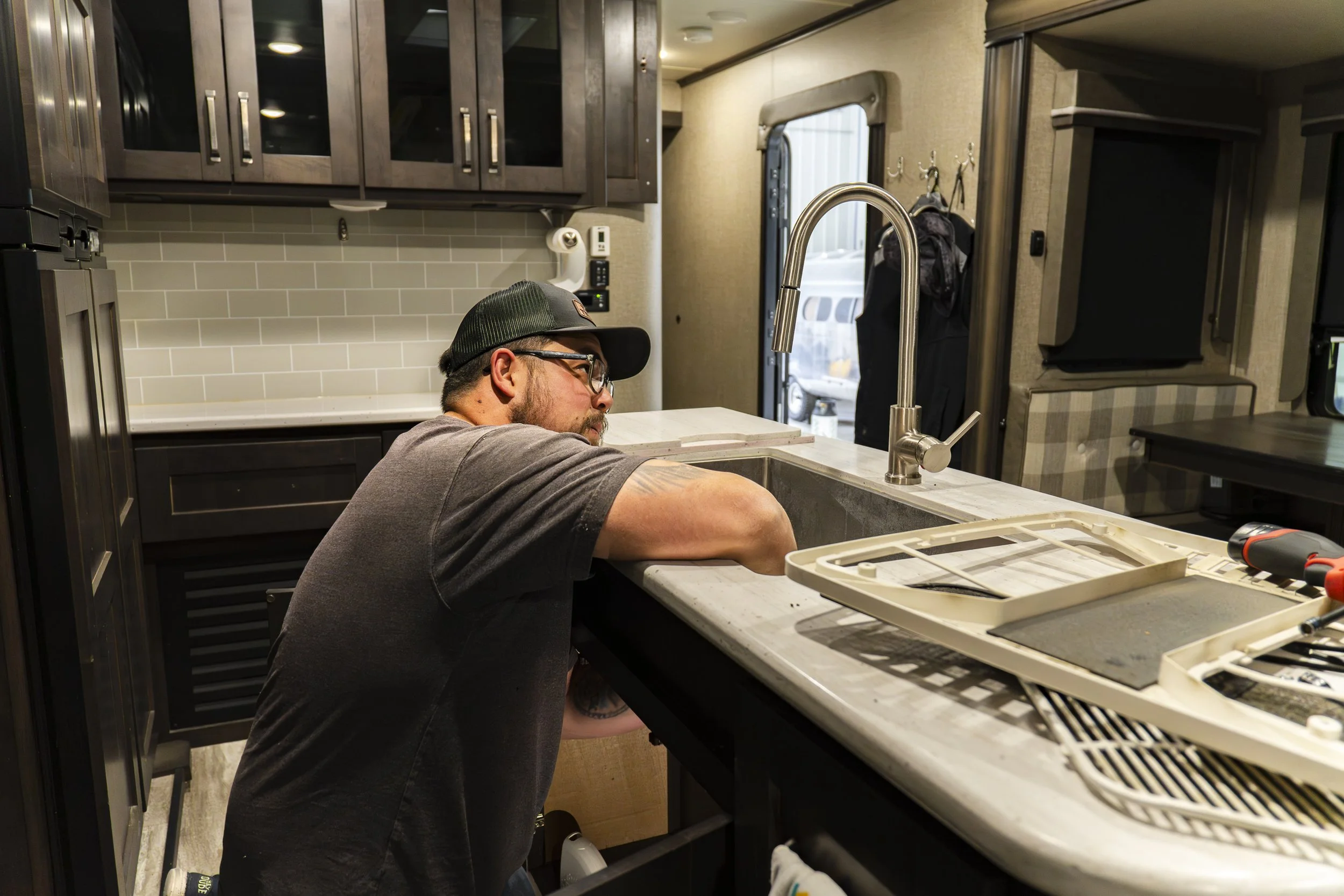 A man wearing glasses, a black cap, and a gray shirt is working on a kitchen sink in a modern RV kitchen, with dark wood cabinets, a tile backsplash, and various tools and parts on the counter.