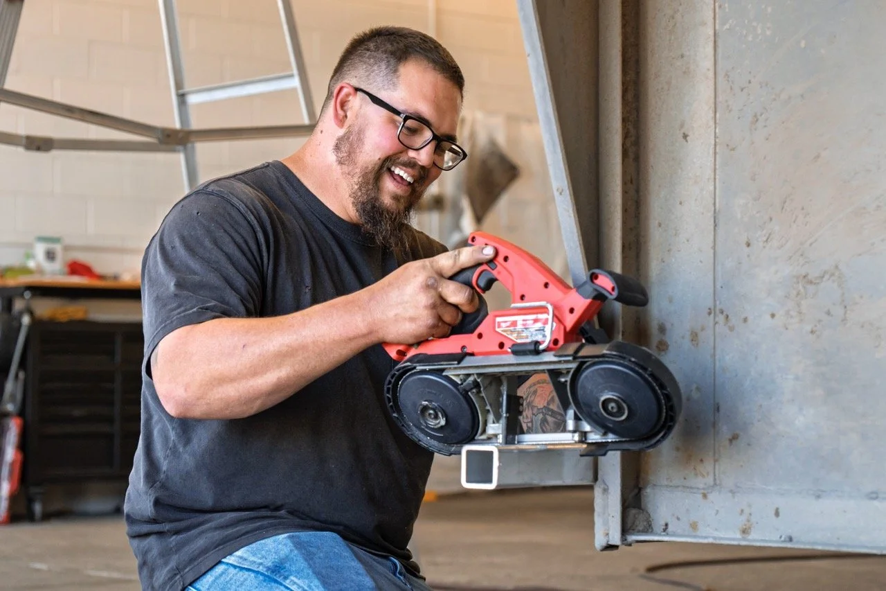 A man with glasses and a beard smiling while using a handheld power saw to cut a metal surface in a workshop.
