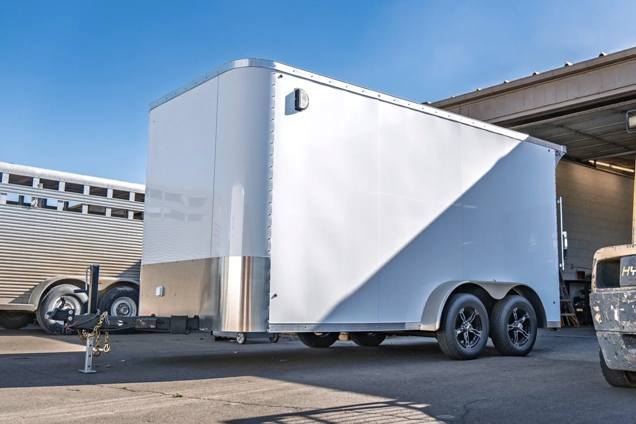 A large, white enclosed trailer with dual axles parked on a paved surface, with another trailer on the left and a building with an open garage in the background.