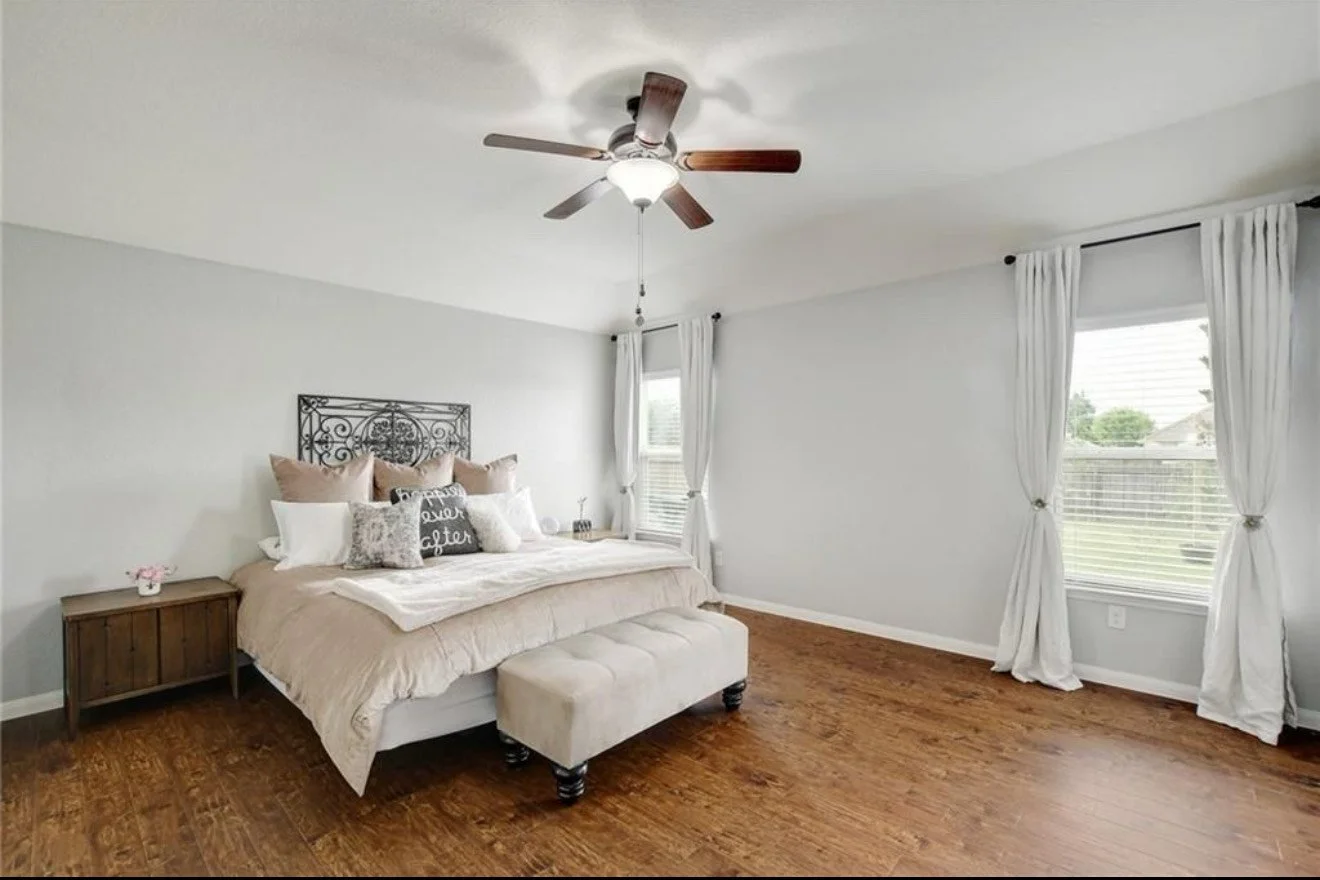 A bedroom with a large bed, beige bedding, and multiple decorative pillows, including a black pillow with white lettering that says 'dream better.' There is a small wooden nightstand with a pink flower arrangement and a white lamp. The room has white walls, hardwood floors, a ceiling fan, and three windows with white curtains. A rectangular beige bench is at the foot of the bed.