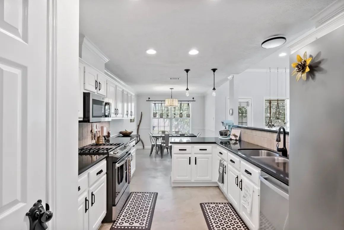 Modern kitchen with white cabinets, black countertops, stainless steel appliances, and a view into the dining area with a window and chandelier.