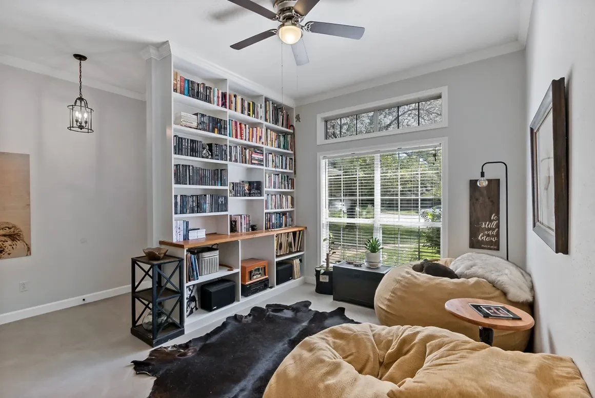 Living room with a bookshelf filled with books, large window with blinds, two beige bean bag chairs, a black rug, a small side table with a magazine, and wall art with a lamp.