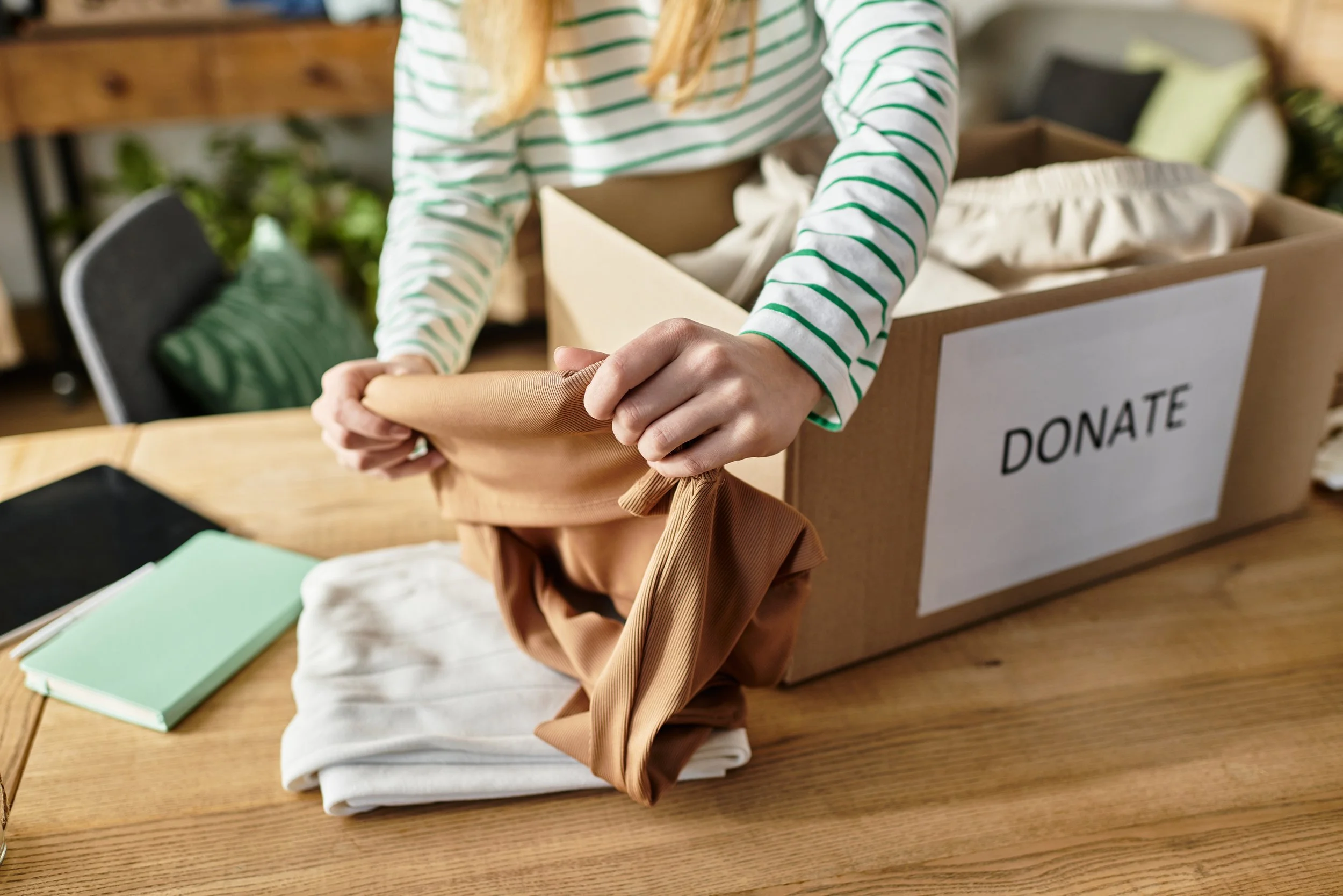 Person packing clothing into a bag for donation, with a box labeled 'DONATE' on a wooden table.