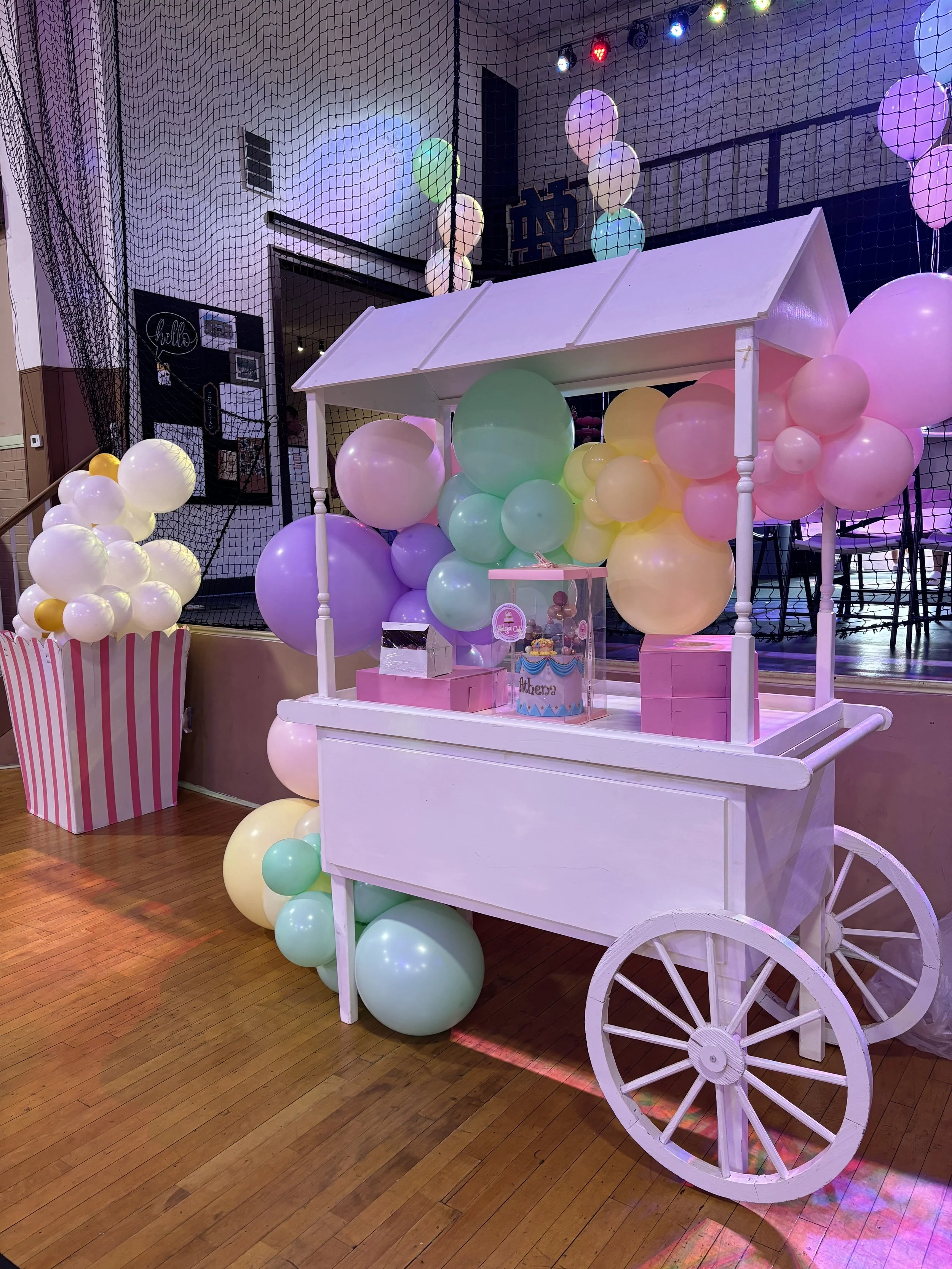 Decorative carnival-style balloon stand with pastel-colored balloons and a small white cart on wooden floor.
