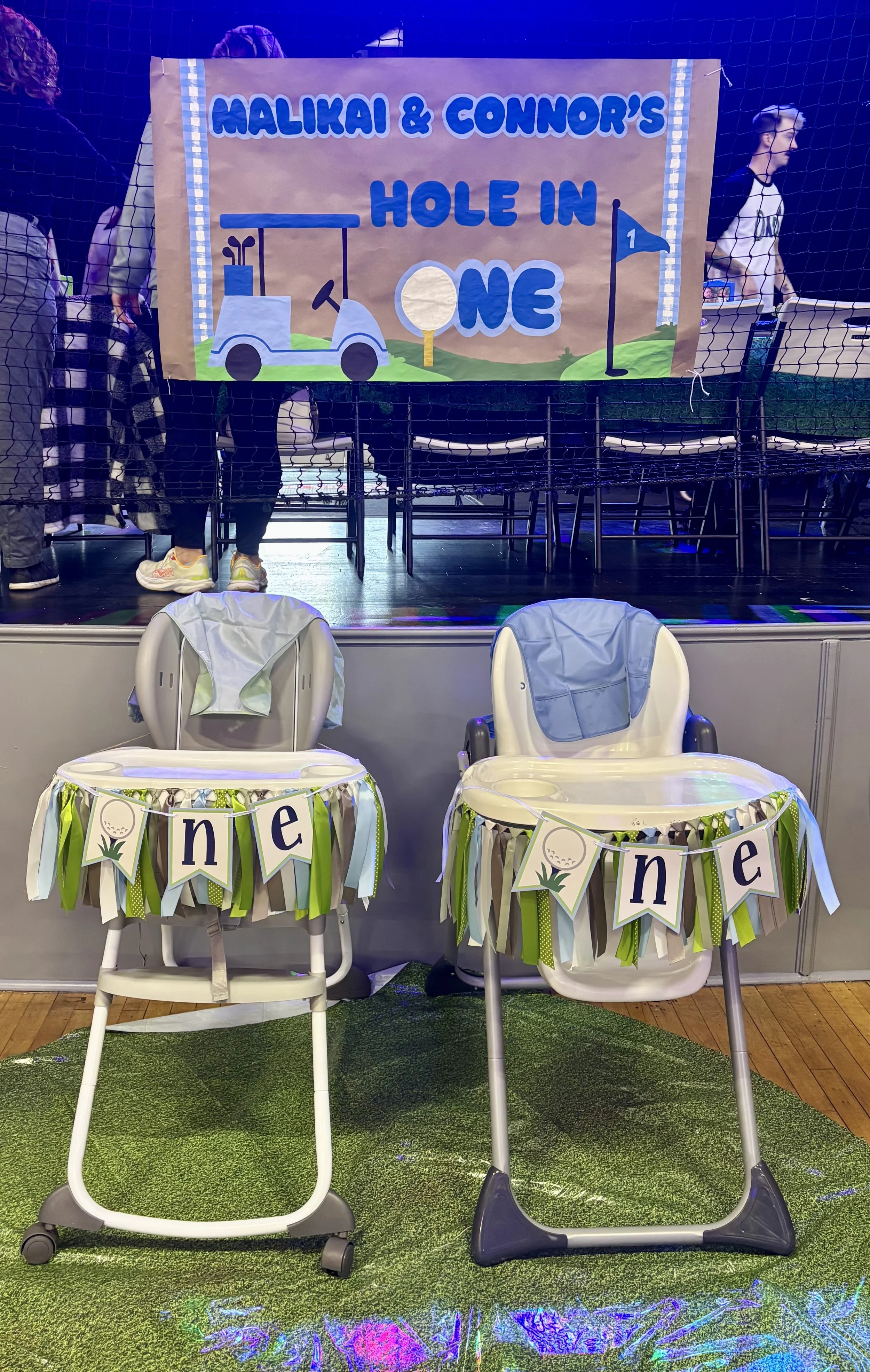 Two decorated high chairs for a children's birthday party with a banner above that reads "Malikai & Connor's Hole in One." The high chairs have green, white, and brown paper decorations and the word "one" on banners attached to them. The background f