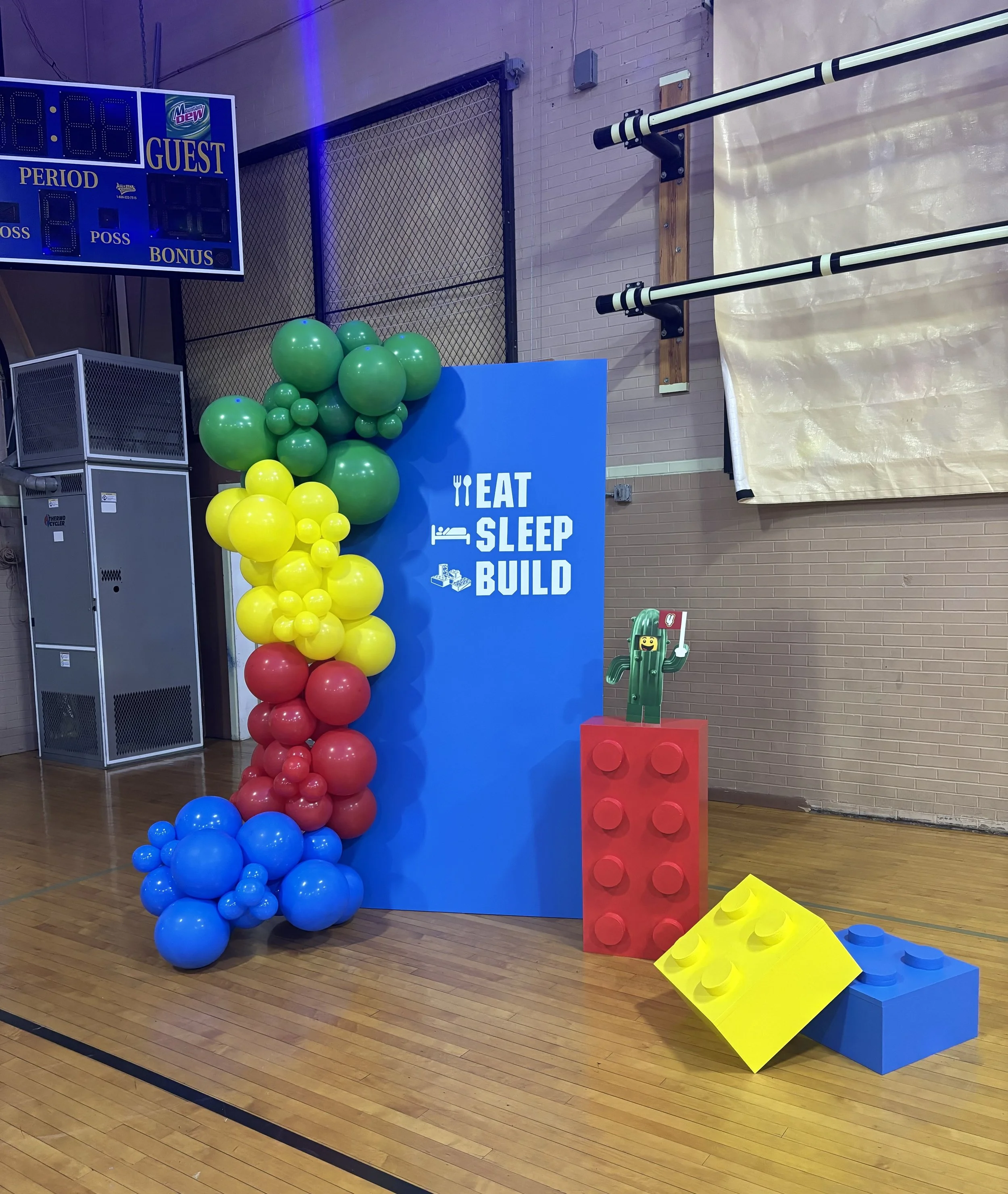 Decorative display in a gymnasium with a blue panel that reads "Eat Sleep Build", surrounded by colorful balloons in blue, red, yellow, and green, and large LEGO-like blocks in yellow, red, and blue.