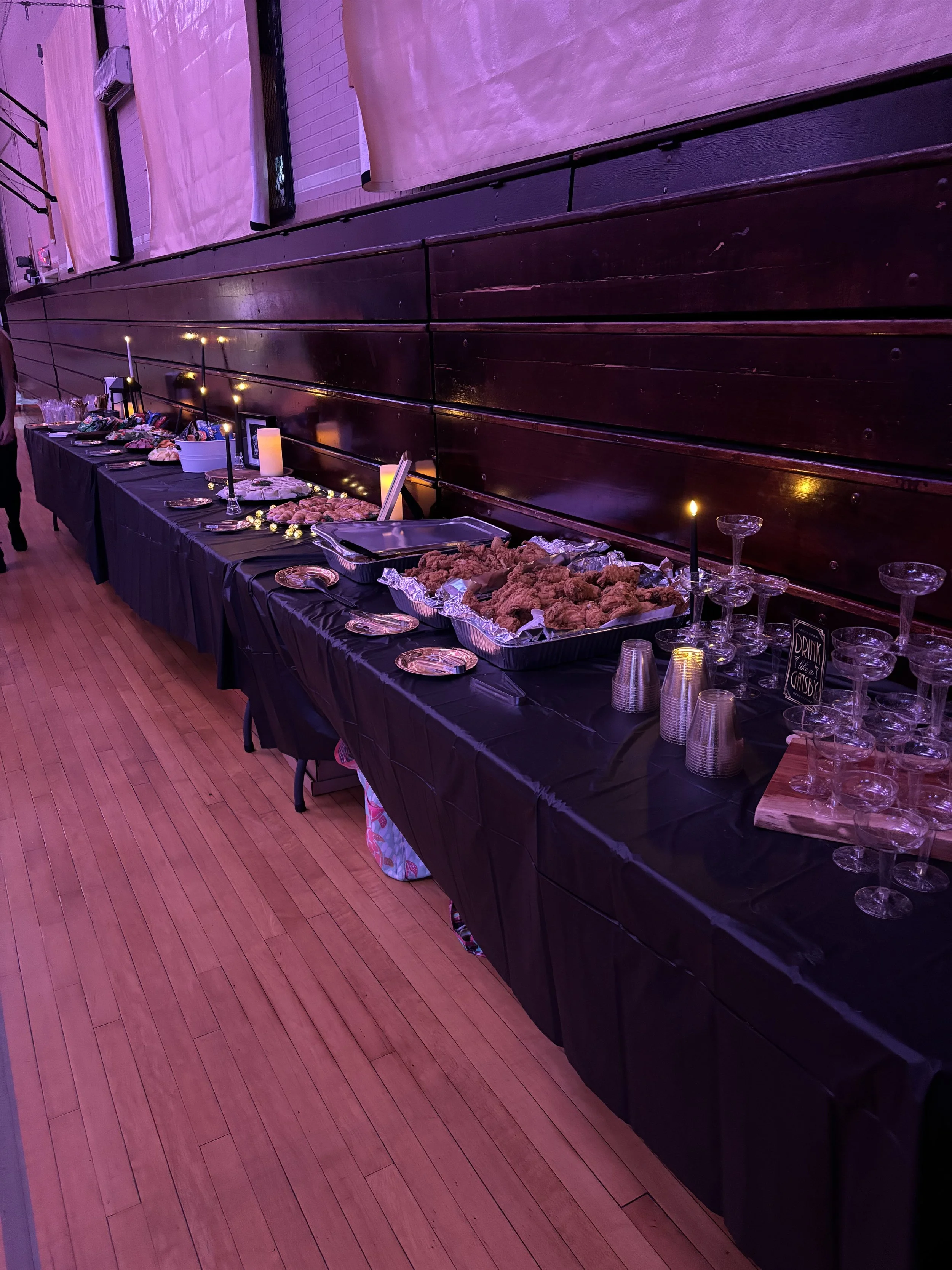 Buffet table with black tablecloth, serving trays of fried chicken, salads, and desserts, decorated with candles and glassware, set against a wooden wall with pink lighting.