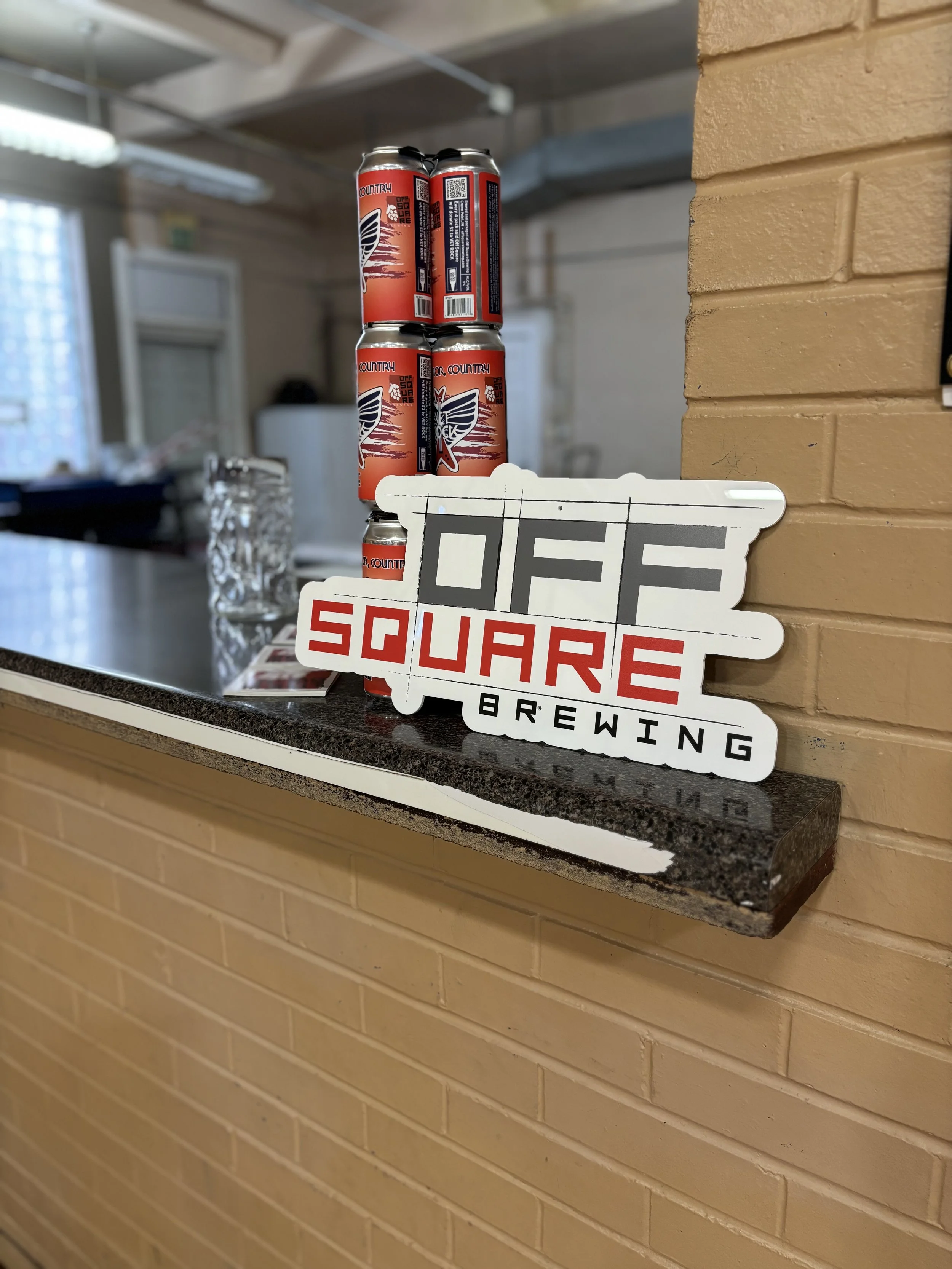 Stack of four cans of beer with a logo featuring wings in front of a brick wall at Off Square Brewing.