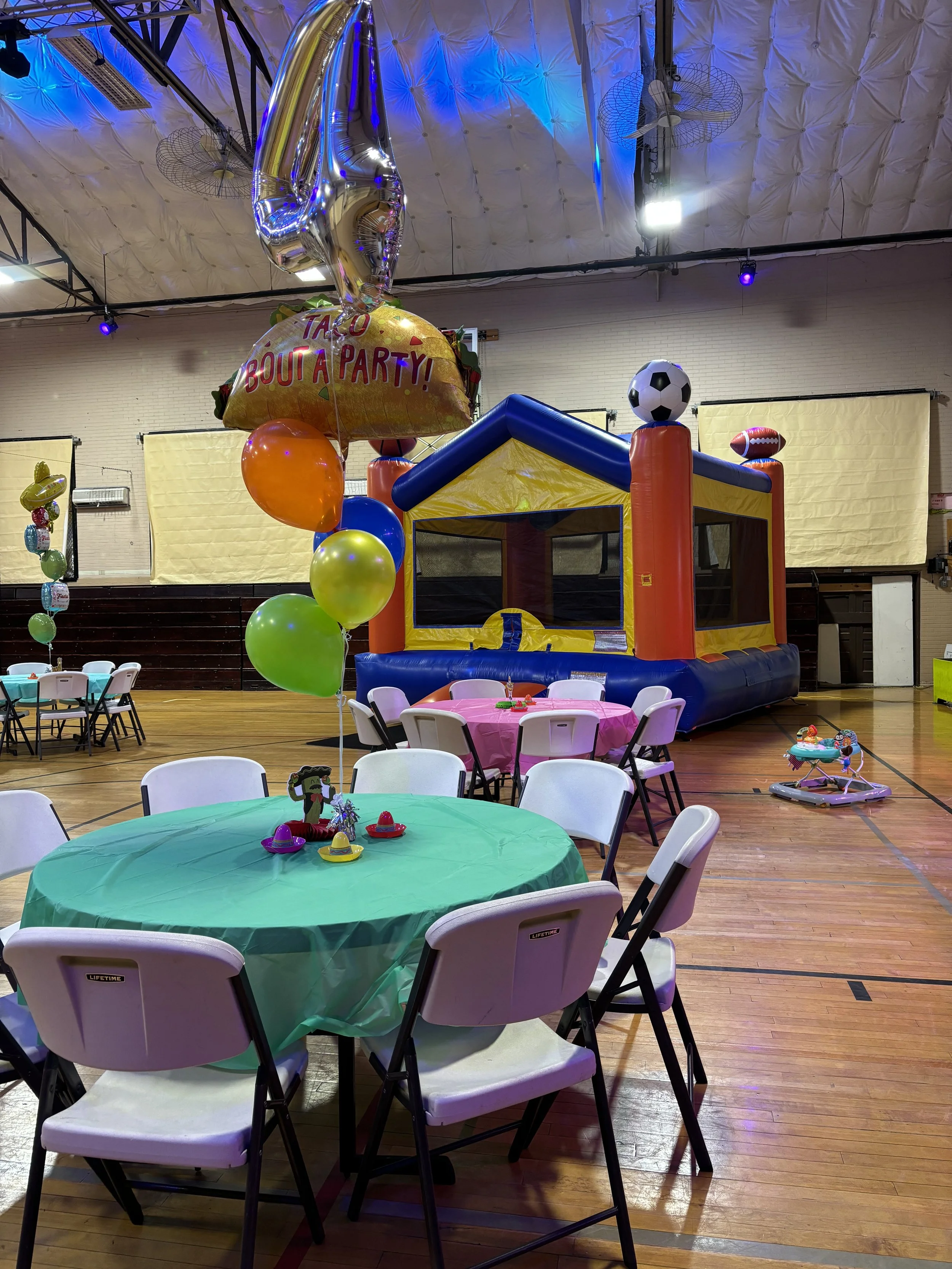 Inside a decorated party venue with tables, chairs, balloons, a bounce house, and a birthday celebration setup.