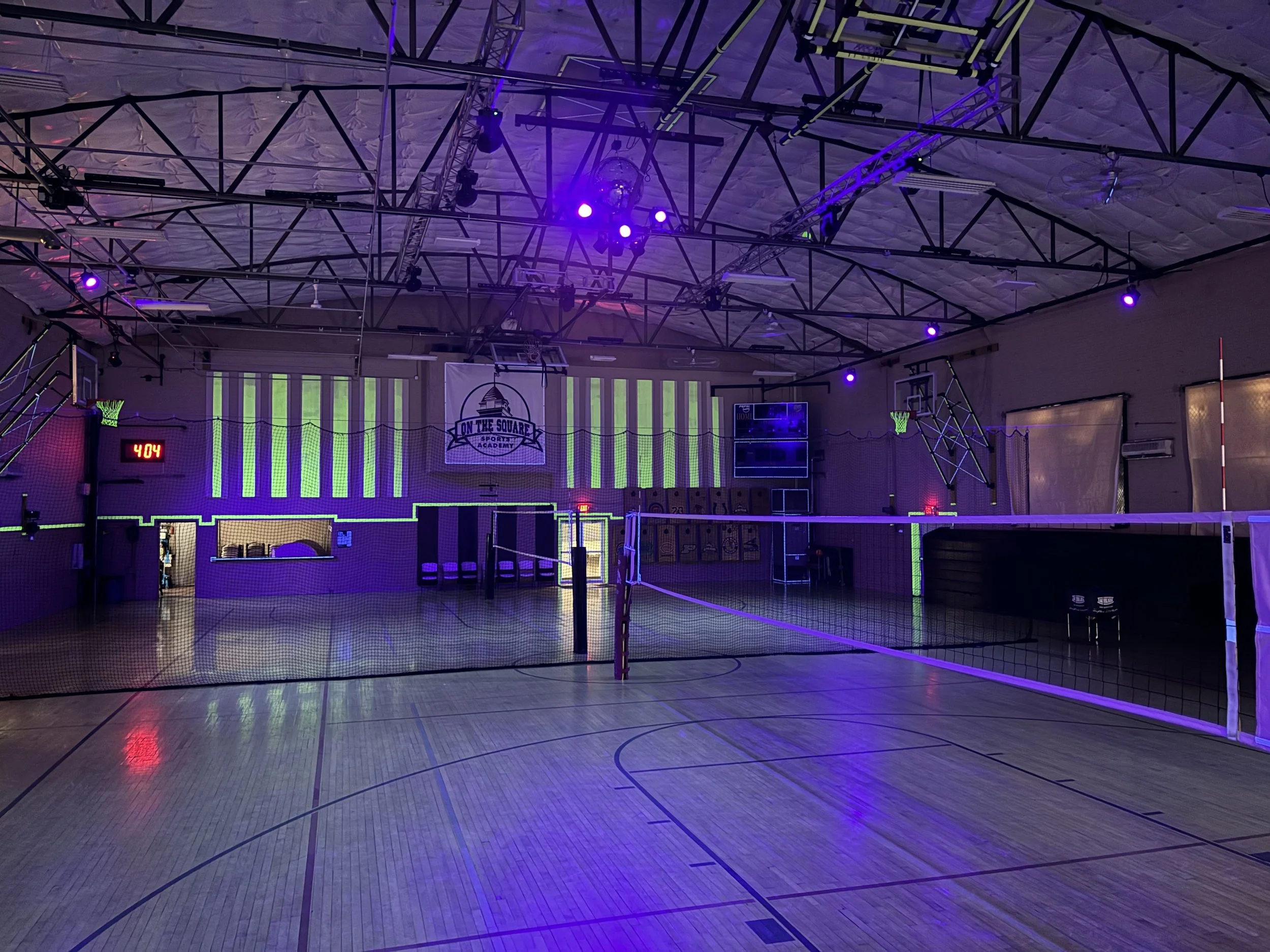 Indoor sports gym with volleyball net, purple and green lighting, a basketball hoop, and a scoreboard on the wall reading 4:04.