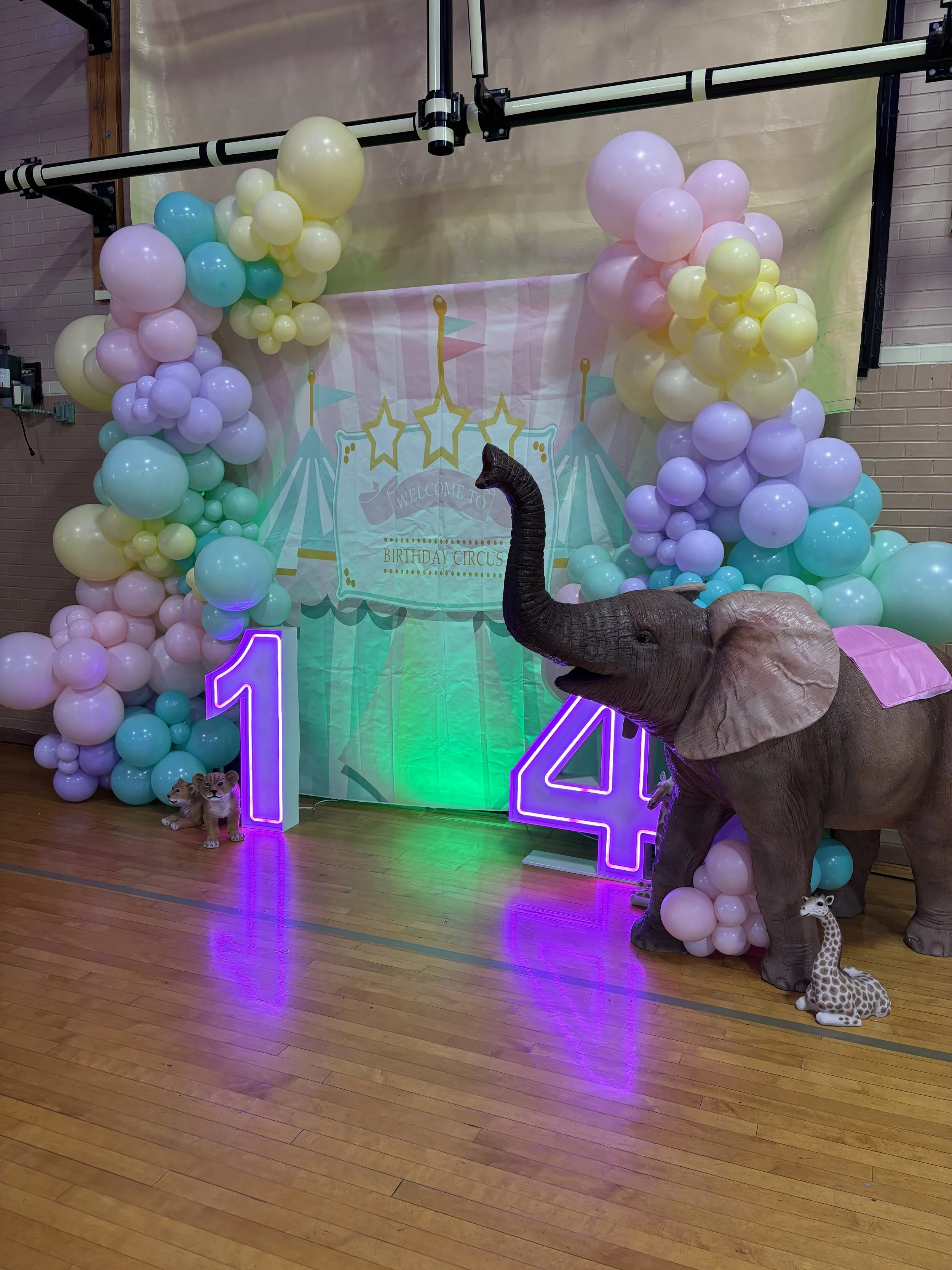 Decorative setup for a child's birthday party featuring a backdrop with circus-themed design, colorful balloon arches in pastel shades, a large inflatable elephant with a pink blanket, neon numbers 1 and 4, and small animal plush toys.