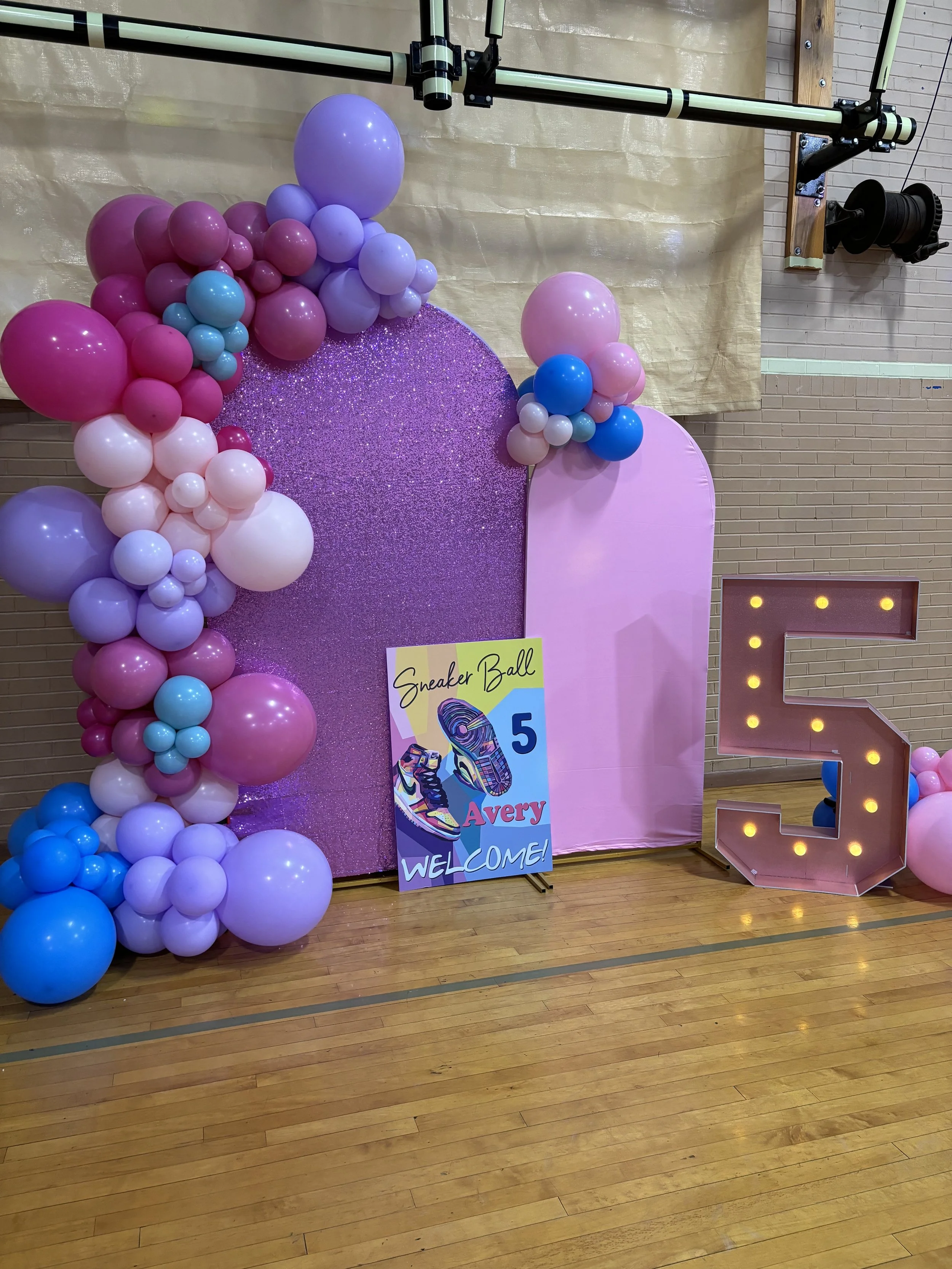 Decorative setup for a birthday celebration with pink, purple, blue, and white balloons arranged on a backdrop, a large illuminated number five, and a sign welcoming guests to a sneaker-themed event for Avery.