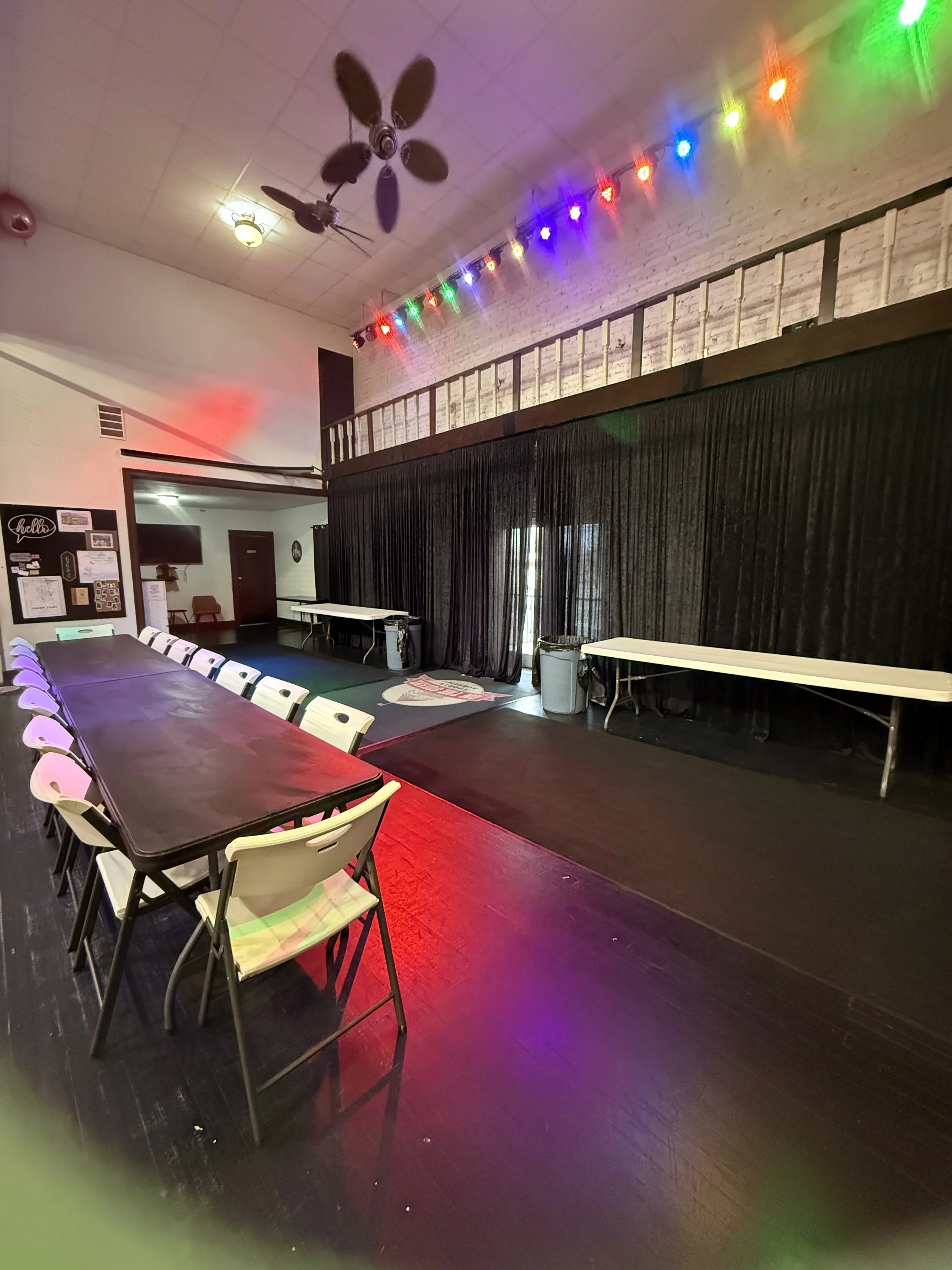 Empty indoor event space with a long table and white chairs, black curtains, multicolored string lights, and a ceiling fan.