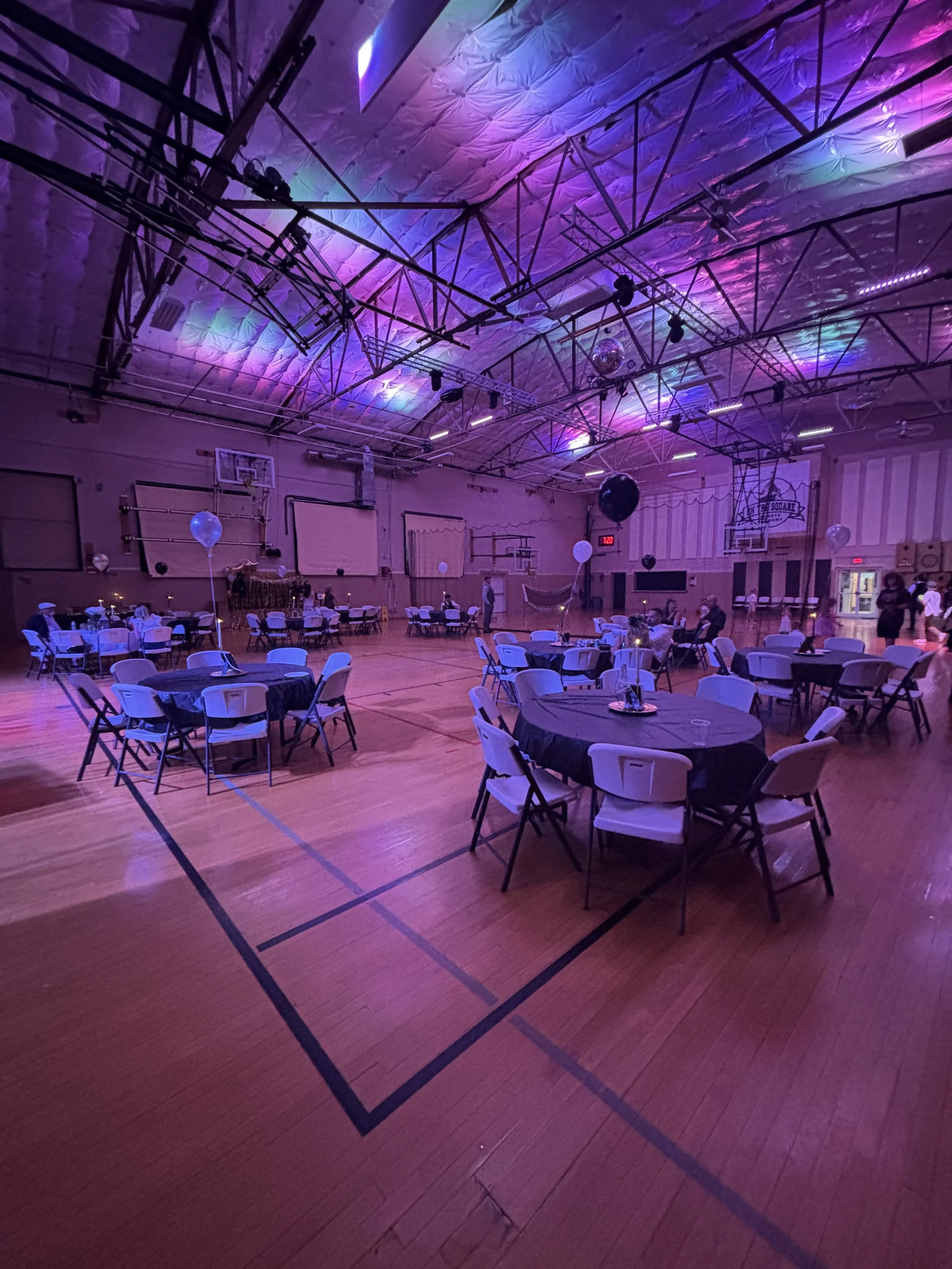 Indoor gymnasium decorated for an event with round tables covered with black tablecloths, chairs, balloons, and purple lighting, with a high ceiling and a basketball hoop.