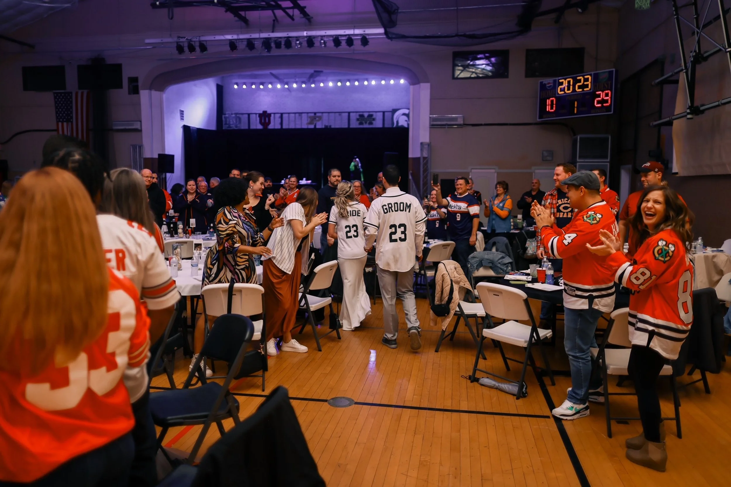 Peopleare gathered in a room, clapping and celebrating at a wedding reception with a sports theme, wearing hockey jerseys, and a couple is walking in the center of the room.