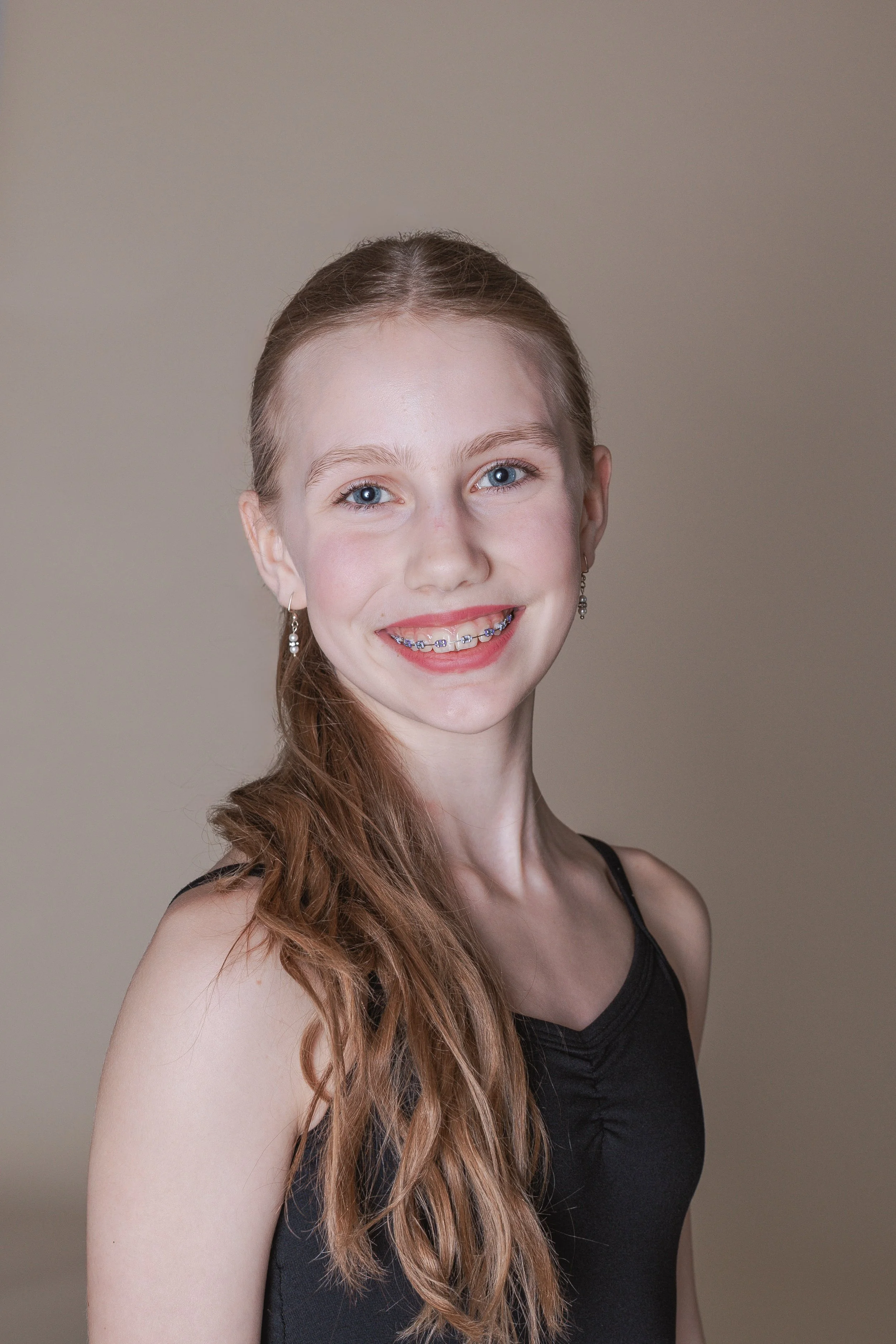 A young girl with long blonde hair, blue eyes, and a big toothy smile, wearing a purple tank top, standing in front of a brick wall.