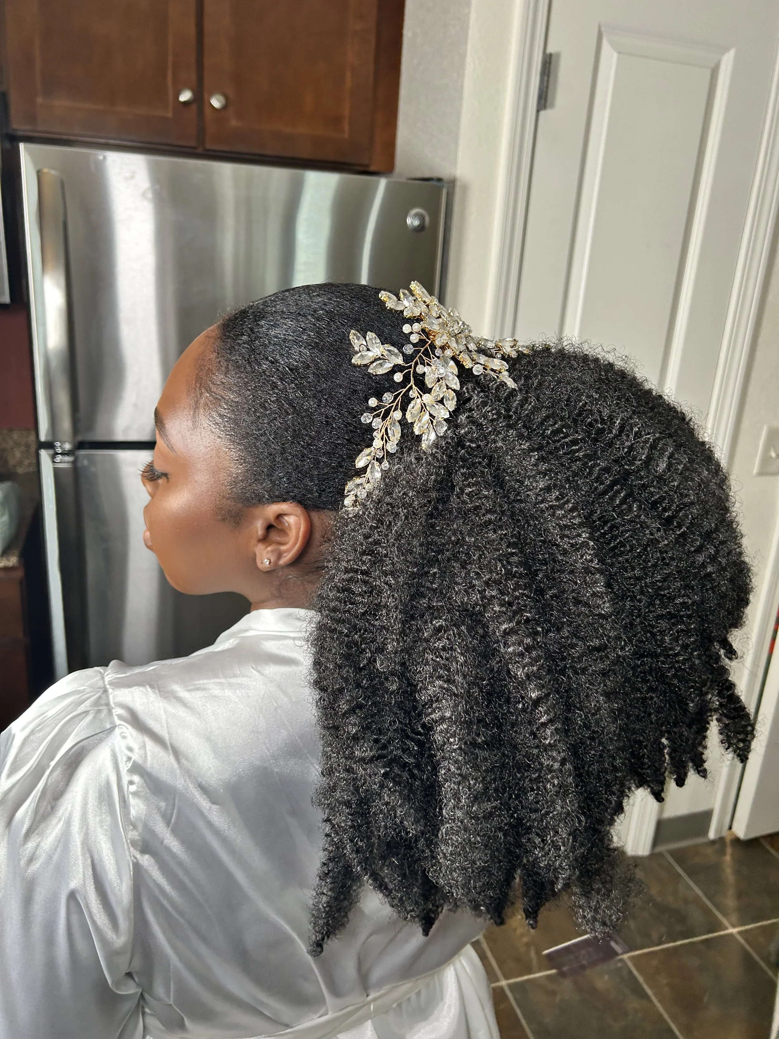 Side view of a woman with curly hair that is styled in an updo and decorated with a jeweled hair accessory. She is wearing a shiny silver satin dress and pearl earrings. The background includes kitchen cabinets and a refrigerator.