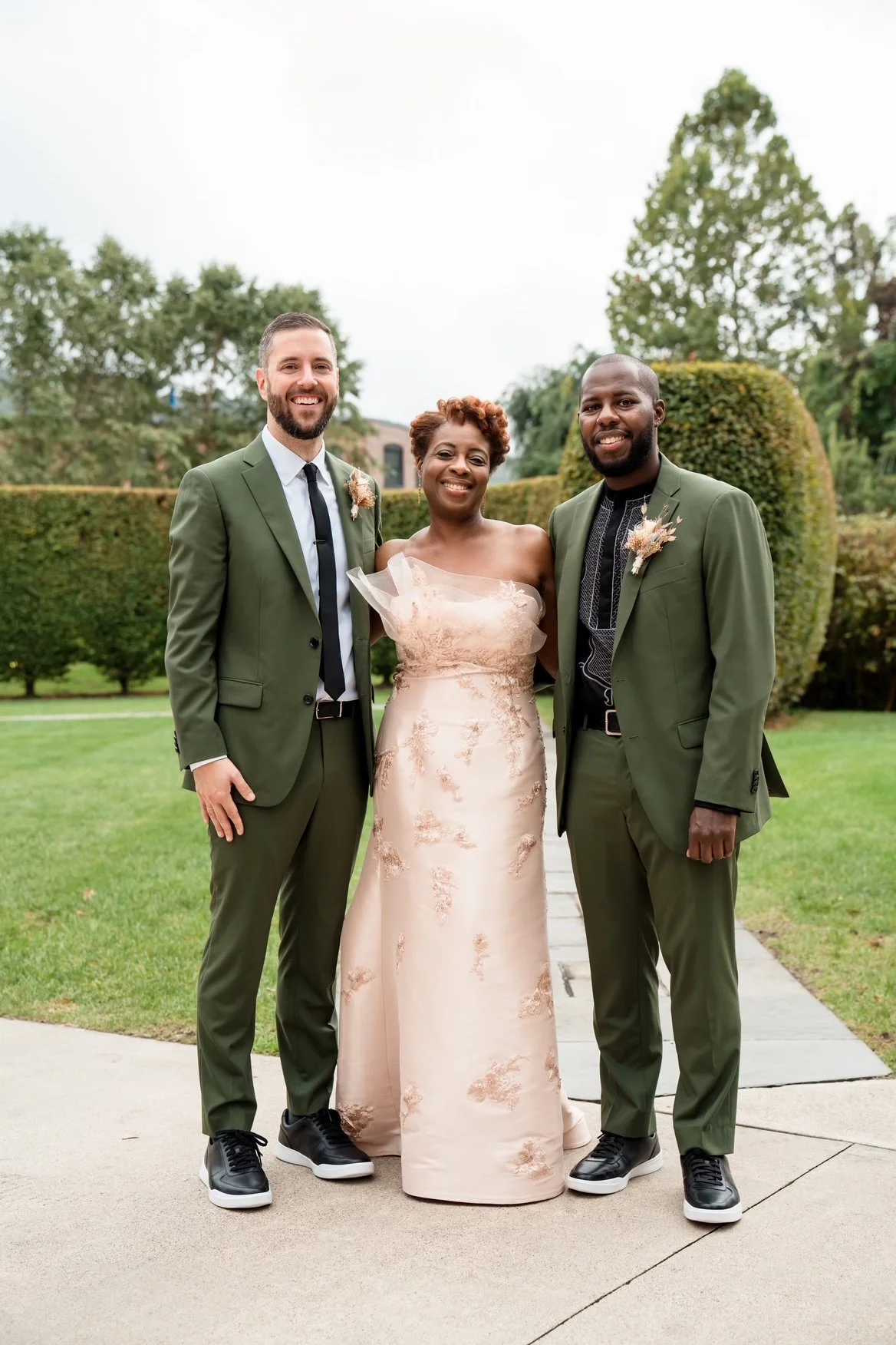 Three people smiling in a park during a wedding celebration. One woman in a beige lace dress and two men in green suits, all holding each other.