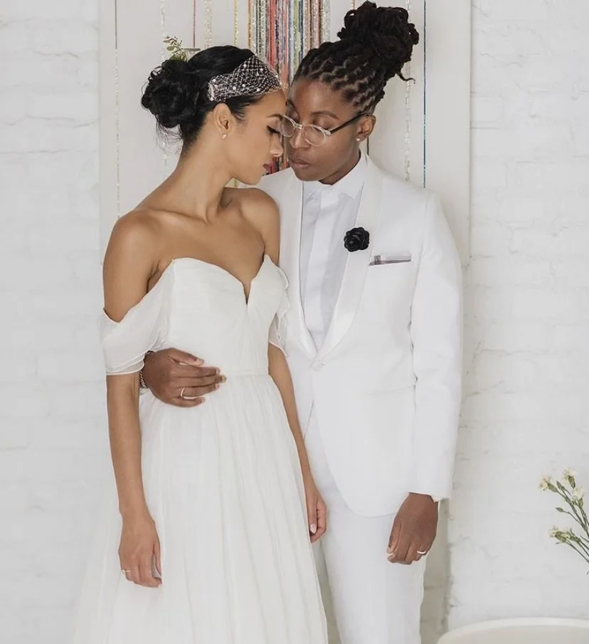 A couple dressed in white wedding attire standing close together with their foreheads touching, in a minimal white room with a bookshelf and a small plant nearby. LBGTQIA Weddings
