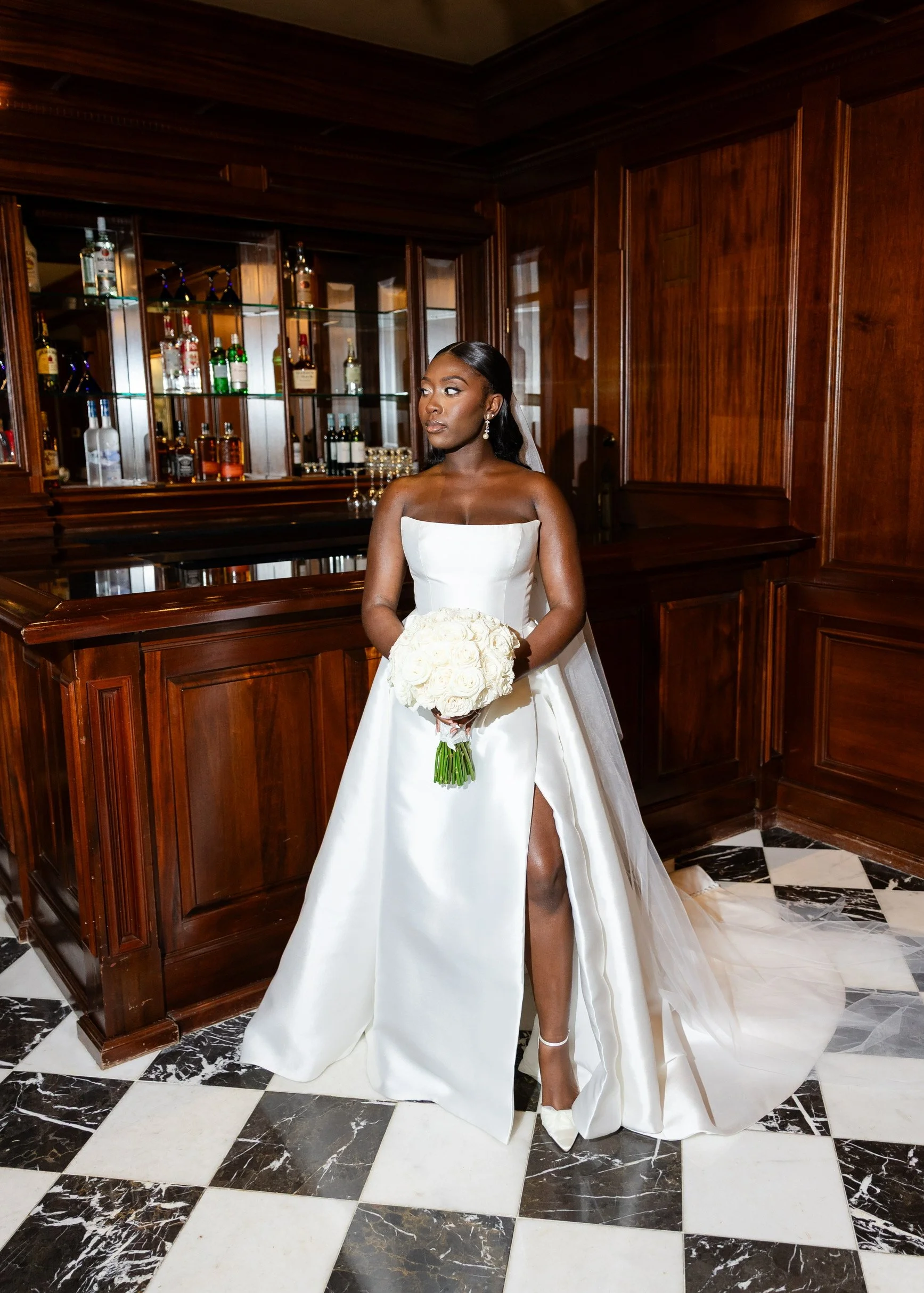A bride in a strapless white wedding gown with a slit, holding a bouquet of white roses, stands in a luxurious wood-paneled room with a black and white checkered marble floor.