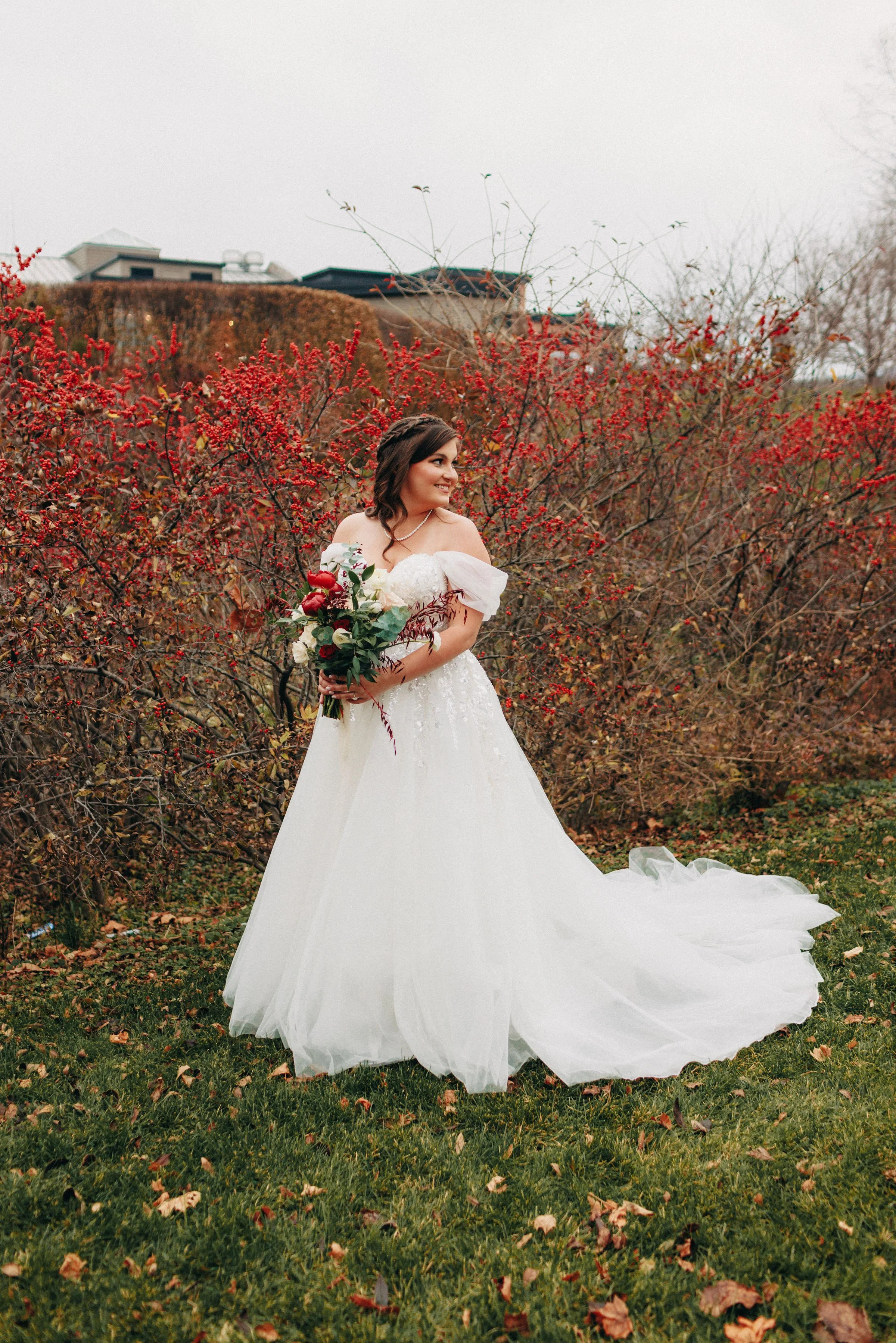 A bride in a white wedding gown holding a bouquet of red, white, and green flowers, standing outdoors among red and brown shrubs and trees with a cloudy sky in the background.