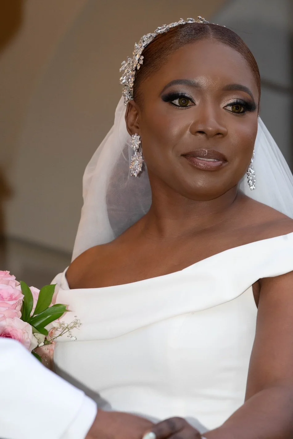 A bride with makeup, jewelry, and a white wedding dress, holding a bouquet of pink roses by Beyond The Knot Collective