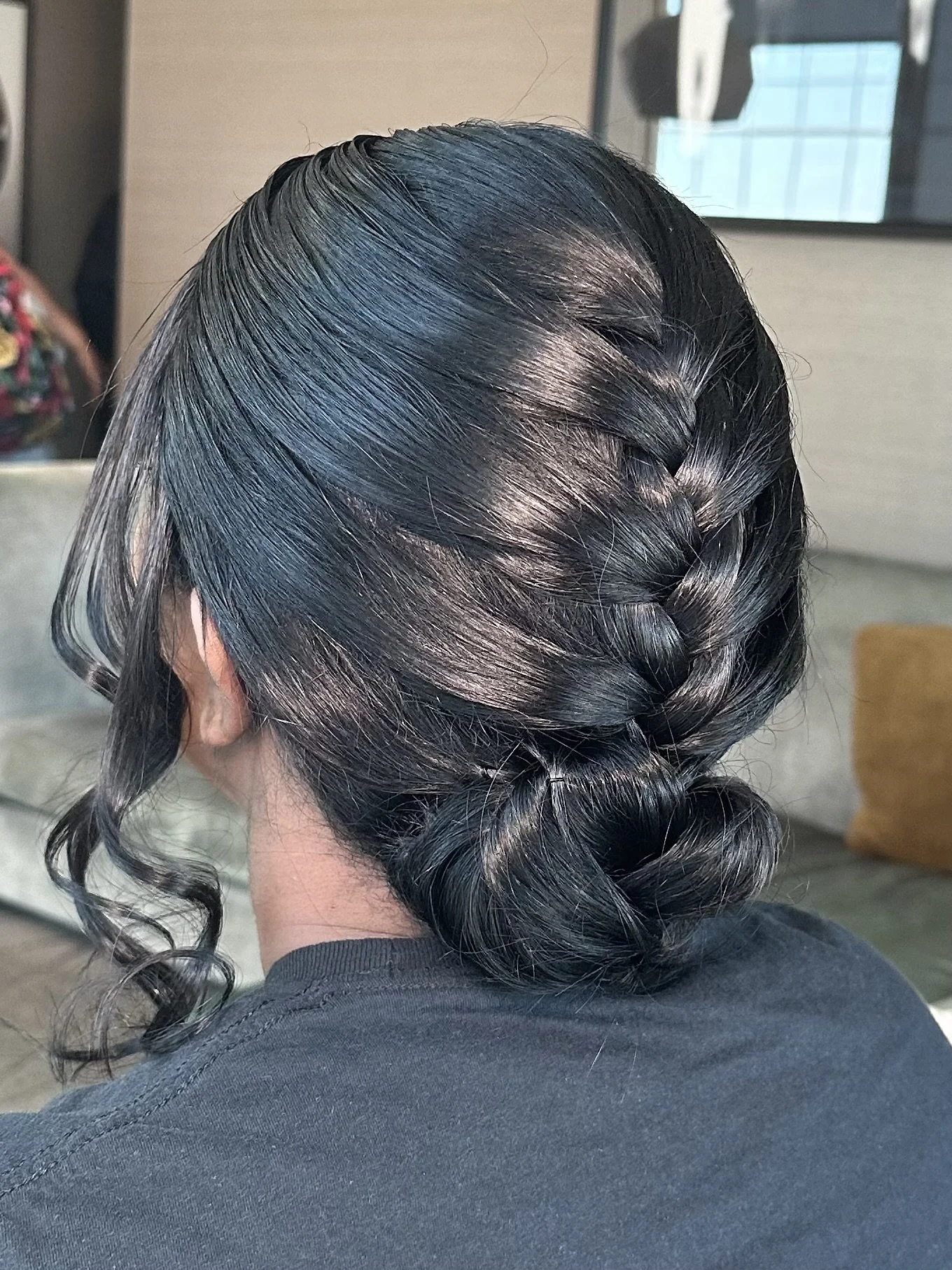 A person's hair styled in a sleek, shiny updo with a braid on the side, with curls hanging down. The background appears to be an indoor setting with a window and a beige wall.