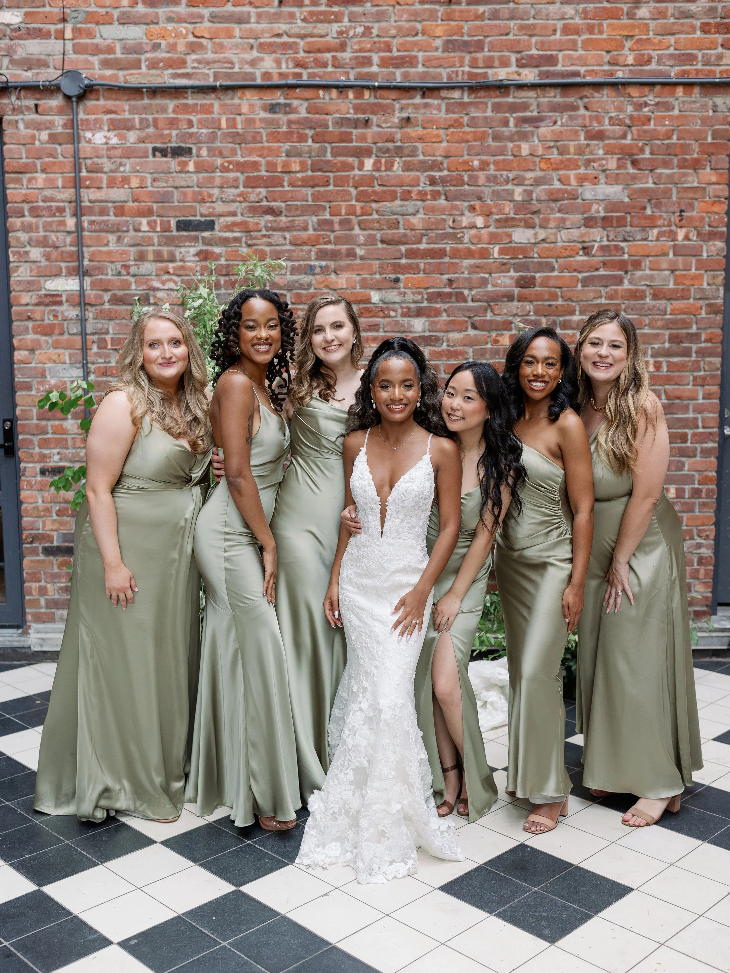 A group of seven women in formal attire, including a bride in a white lace gown and six bridesmaids in matching sage green dresses, standing in front of a brick wall with a black and white checkered floor.
