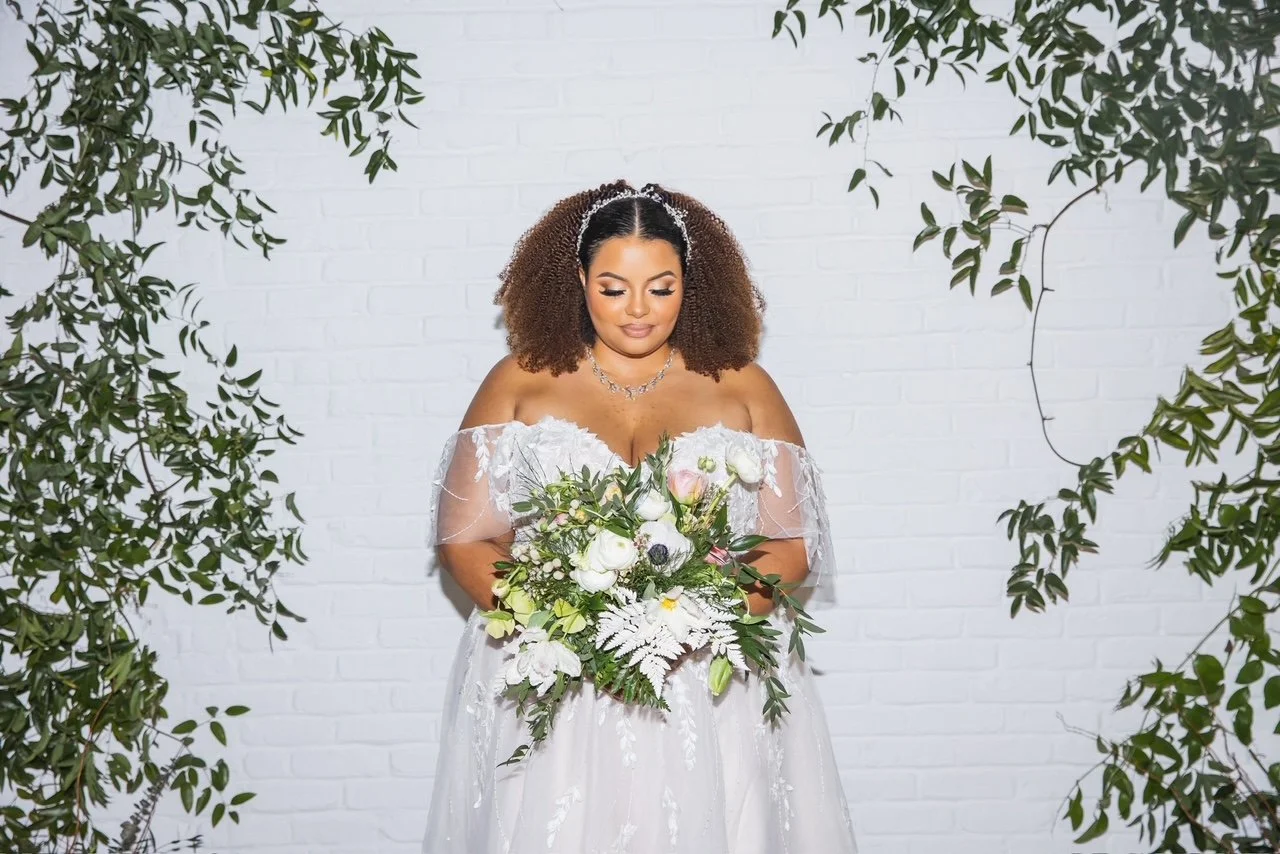 A bride wearing an off-the-shoulder white wedding gown holding a bouquet of white and pink flowers, standing against a white brick wall surrounded by green foliage.