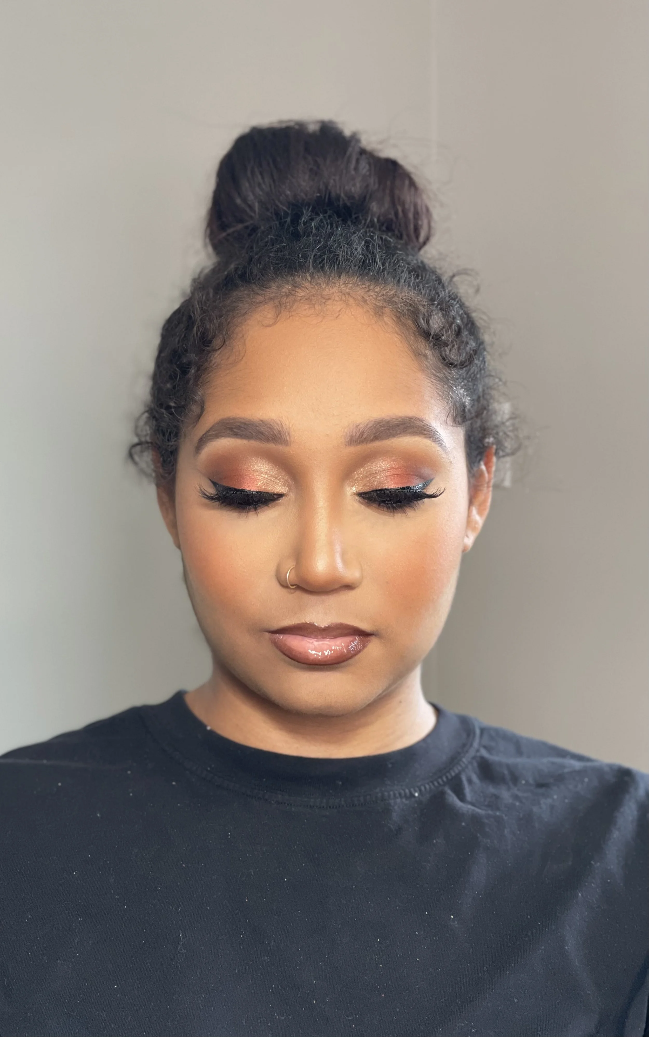 A woman with her eyes closed, makeup including shimmery eyeshadow, eyeliner, and glossy lips, her hair styled in a high bun with some curly strands, wearing a black top with small white specks, standing against a plain gray background.