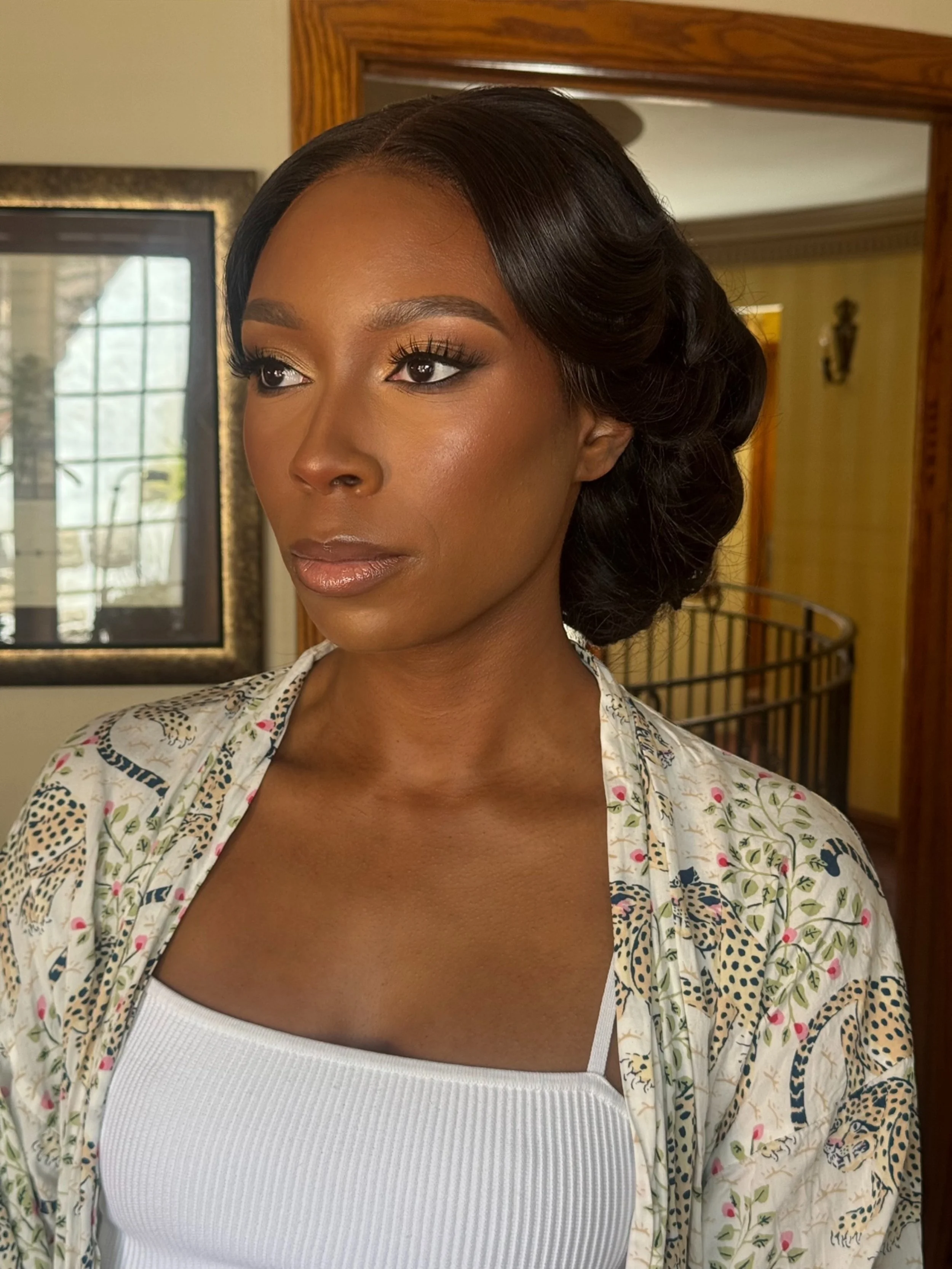 A woman with dark hair styled in waves, wearing makeup, a white ribbed tank top, and a floral patterned blazer standing indoors near a mirror and window.