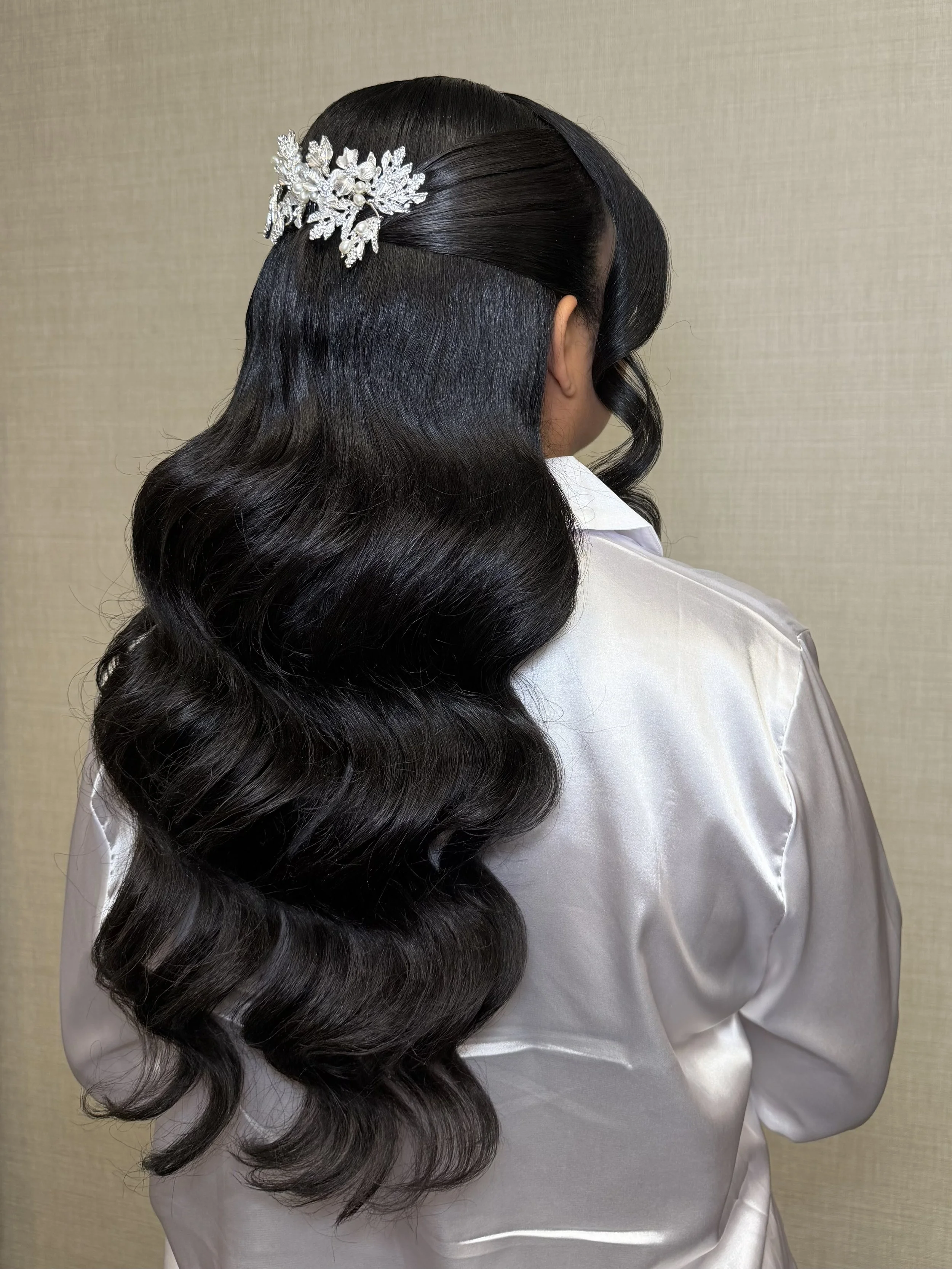 A woman with styled, long, dark, wavy hair wearing a decorative silver hairpiece with flowers and leaves, dressed in a silky white garment.