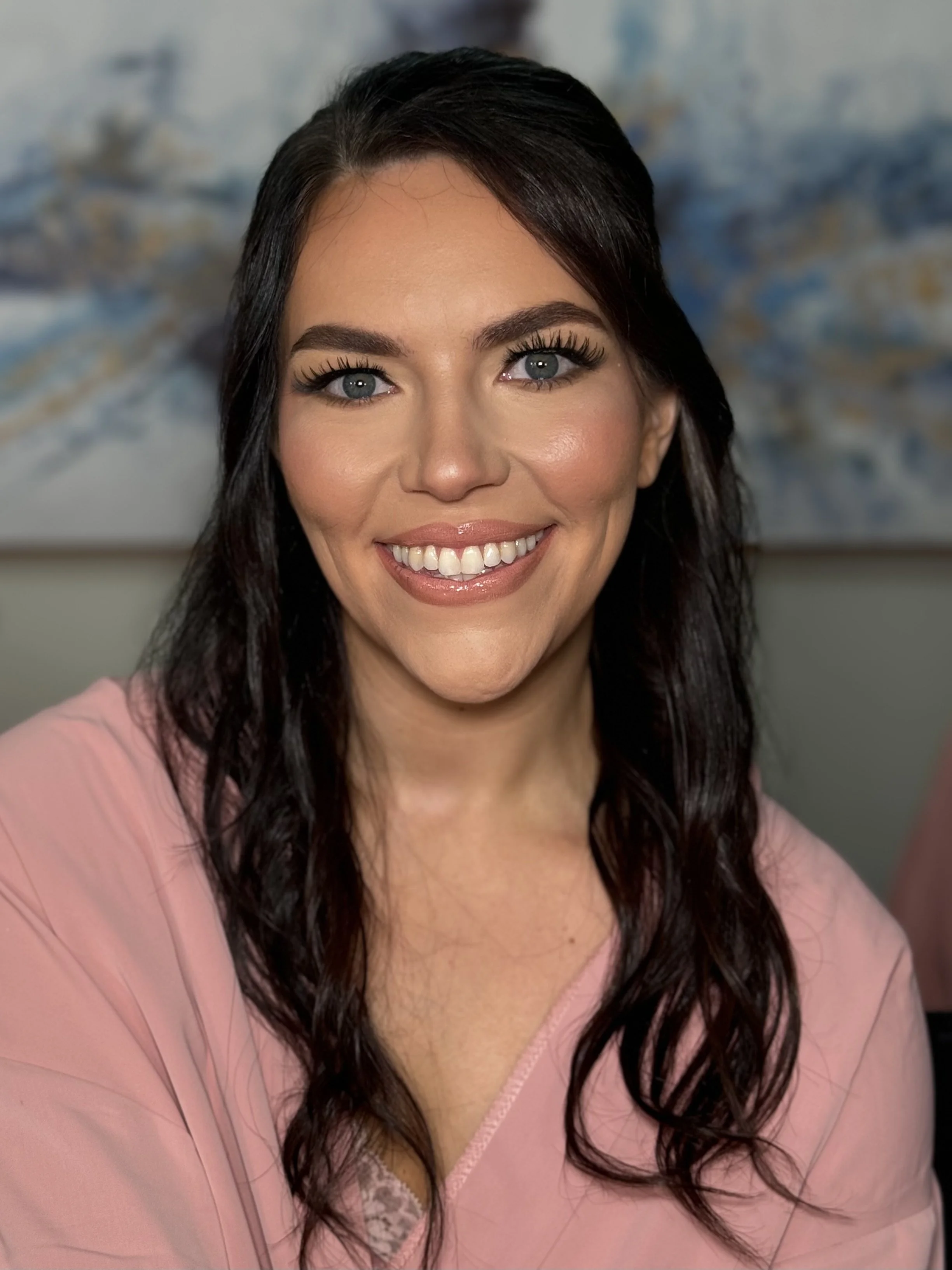A woman with long, dark, wavy hair, wearing a pink top, smiling showing her teeth, with some makeup, standing in front of a textured blue and white background.