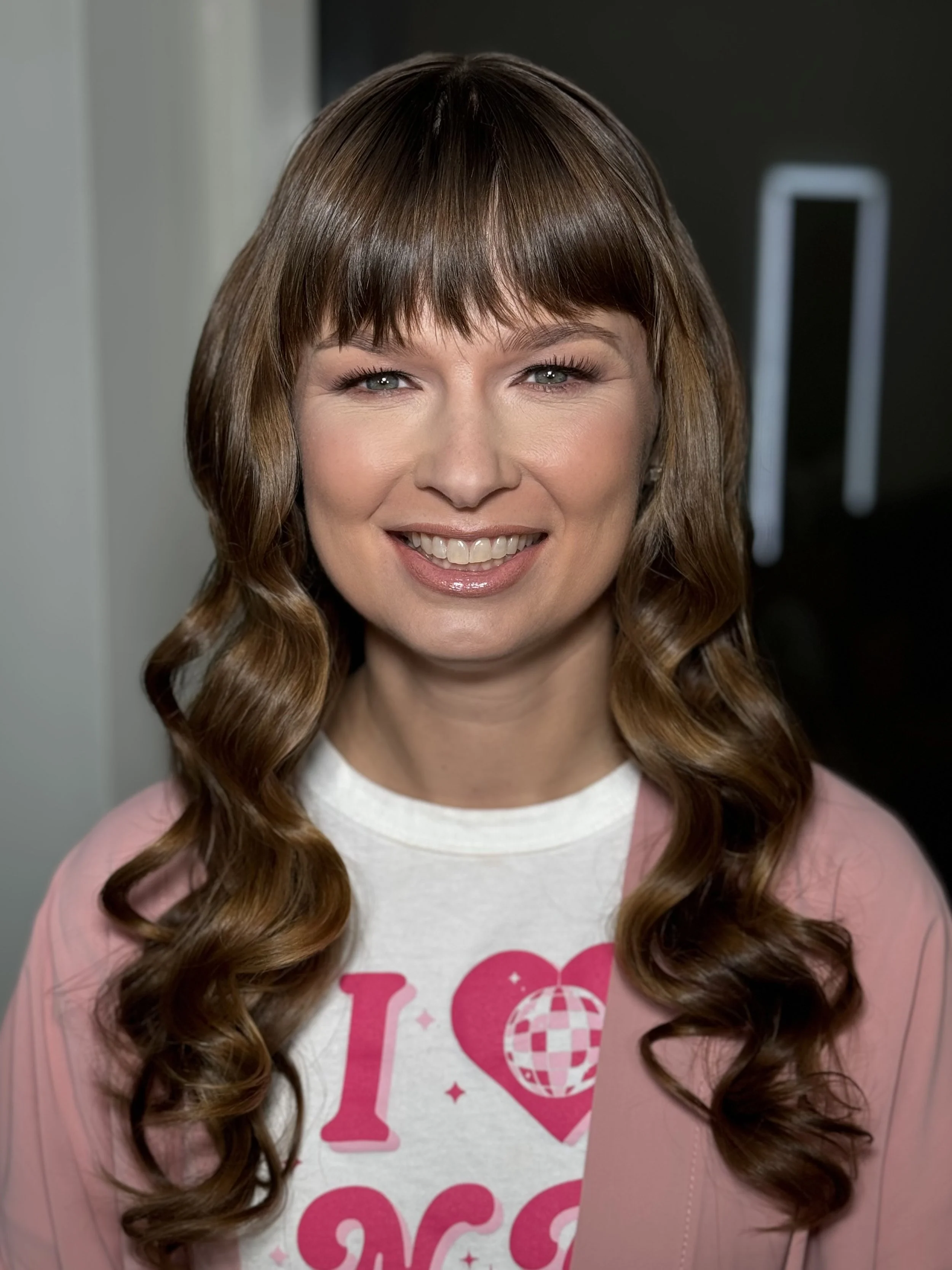 A smiling woman with long, curly brown hair and light skin, wearing a white t-shirt with pink print and a light pink cardigan.