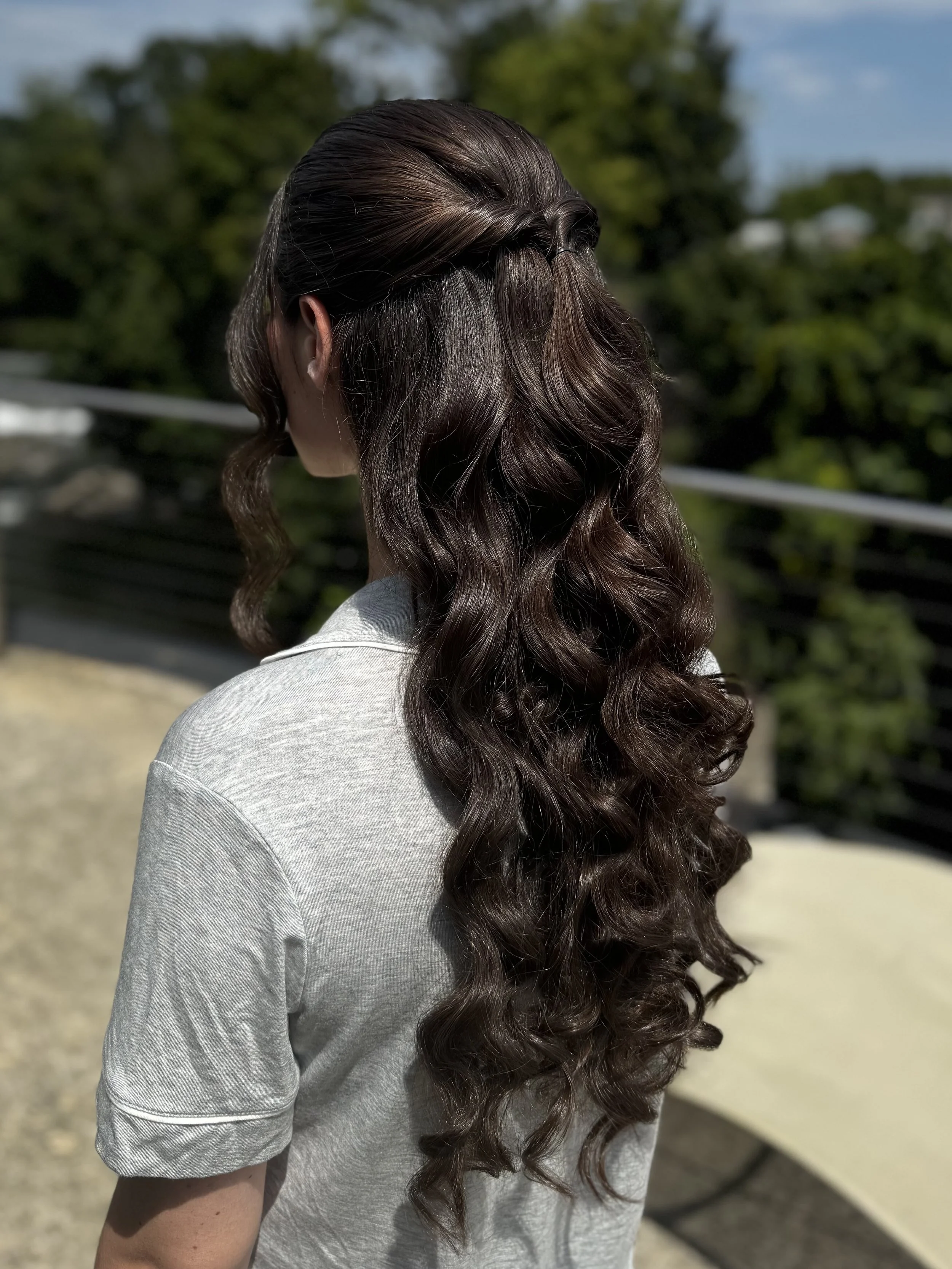 A woman with long, dark, wavy hair wearing a gray shirt standing outdoors near a dark railing with greenery in the background.