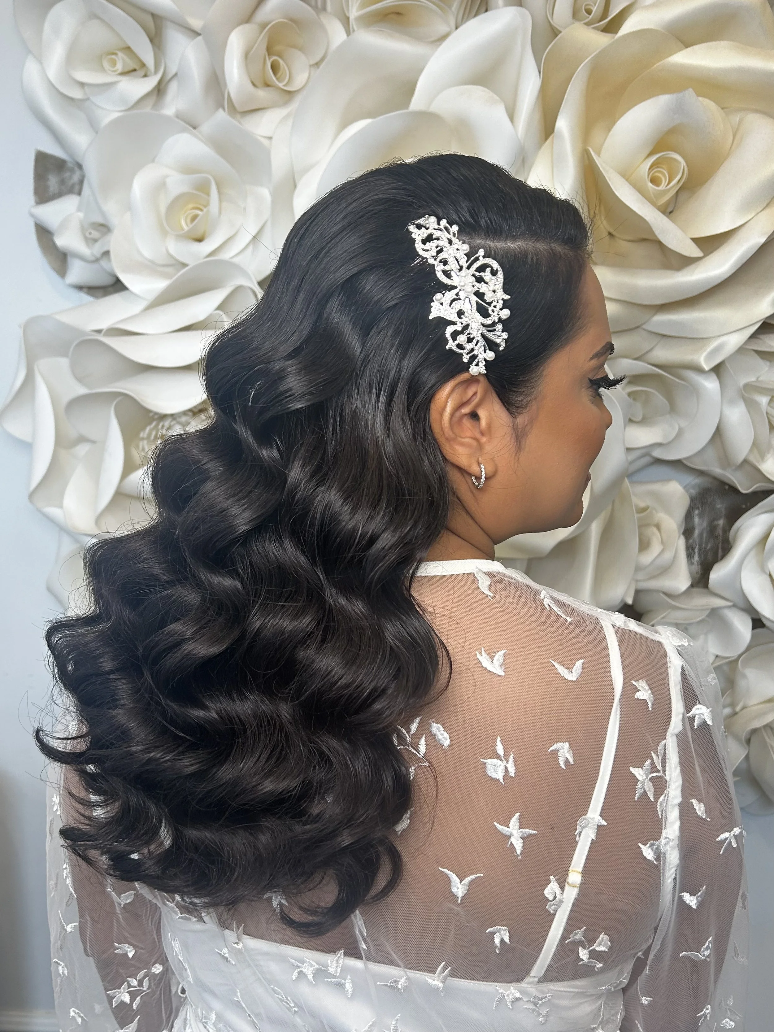 A woman with long, dark, wavy hair wearing a decorative hair clip adorned with crystals, standing in front of a backdrop made of white artificial roses.