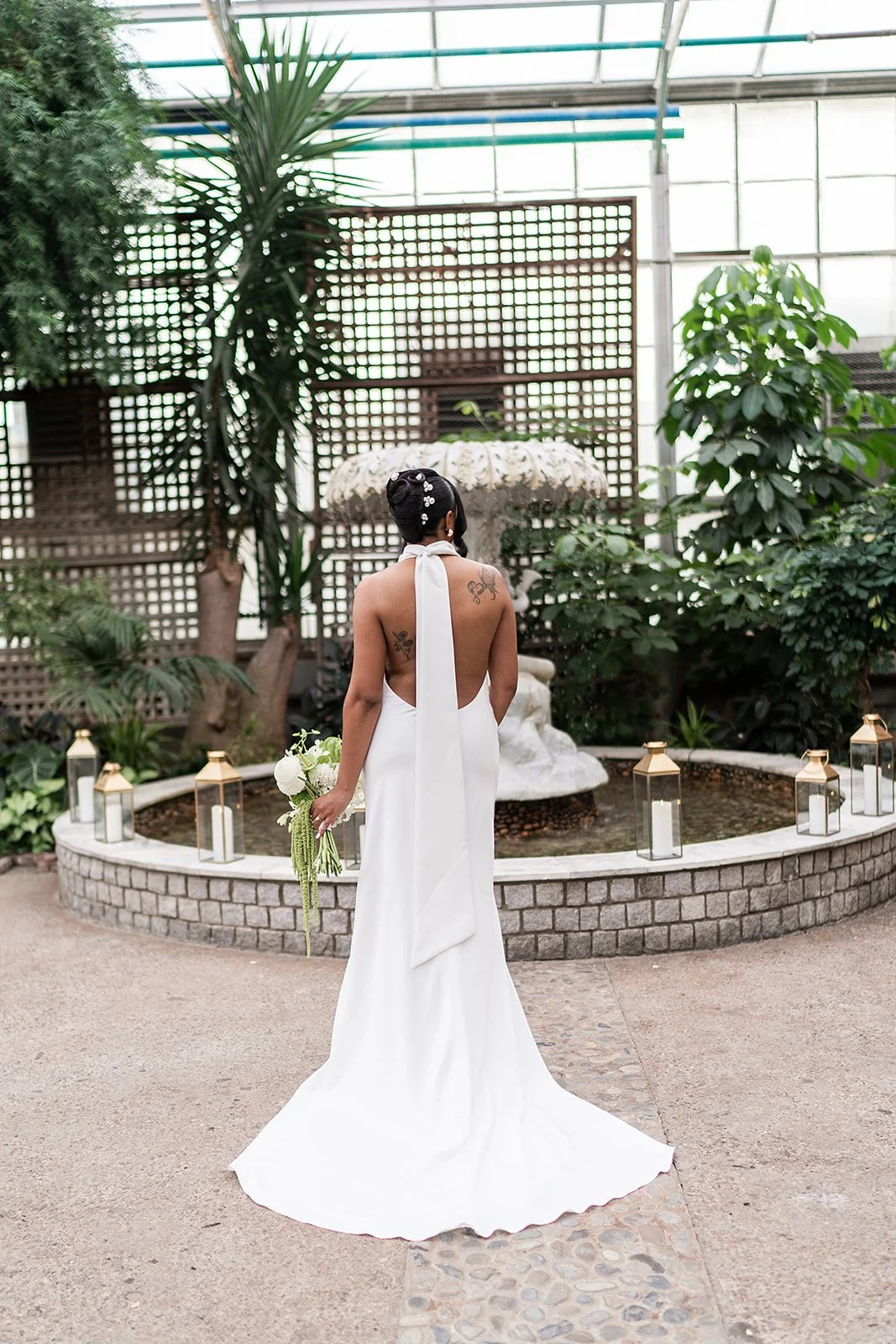 Bride in a white gown holding a bouquet of white flowers, standing in a greenhouse with green plants, a fountain, and lanterns.