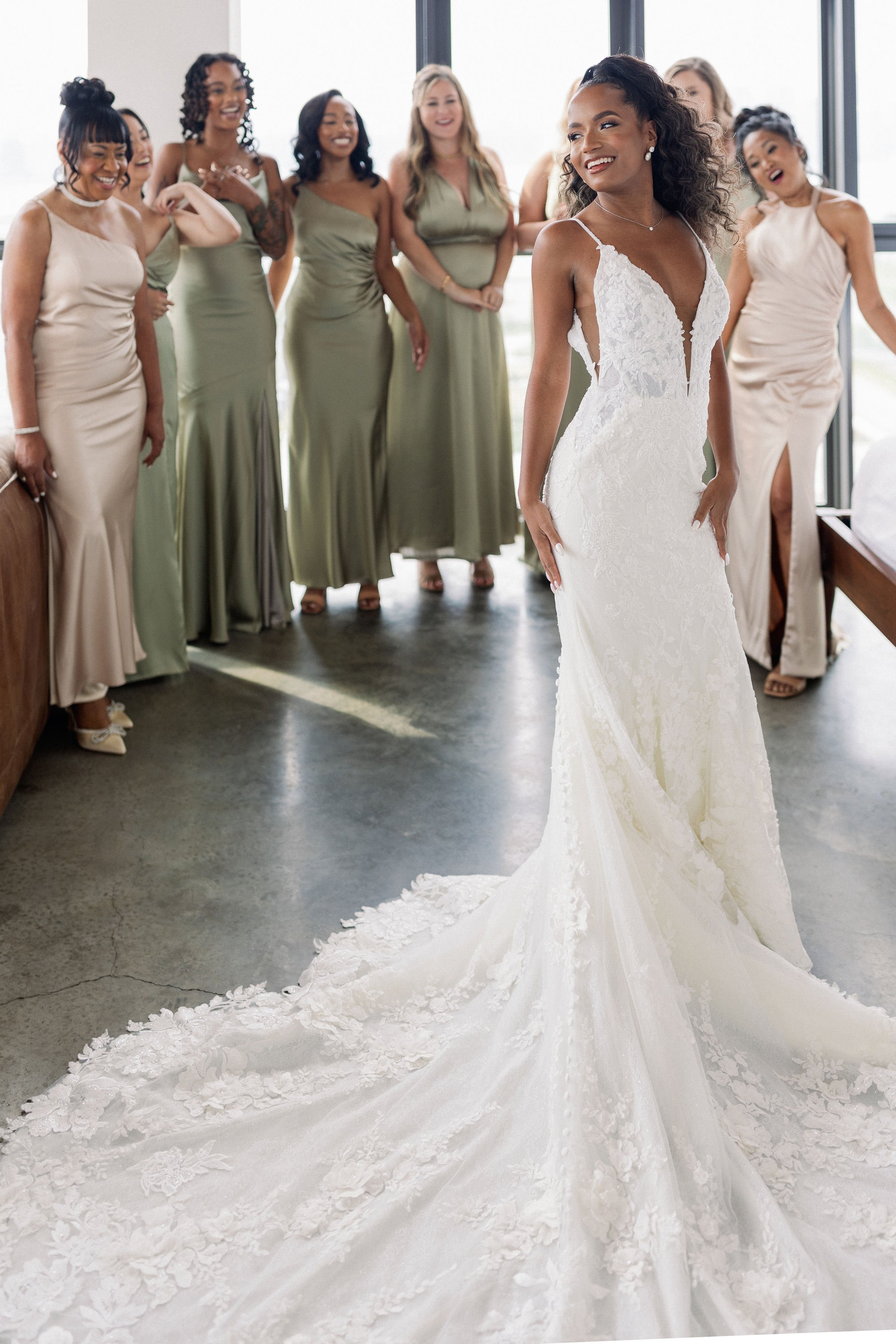 A bride in a white lace wedding dress with a train, standing in front of a group of women in matching green bridesmaid dresses, inside a modern building with large windows.