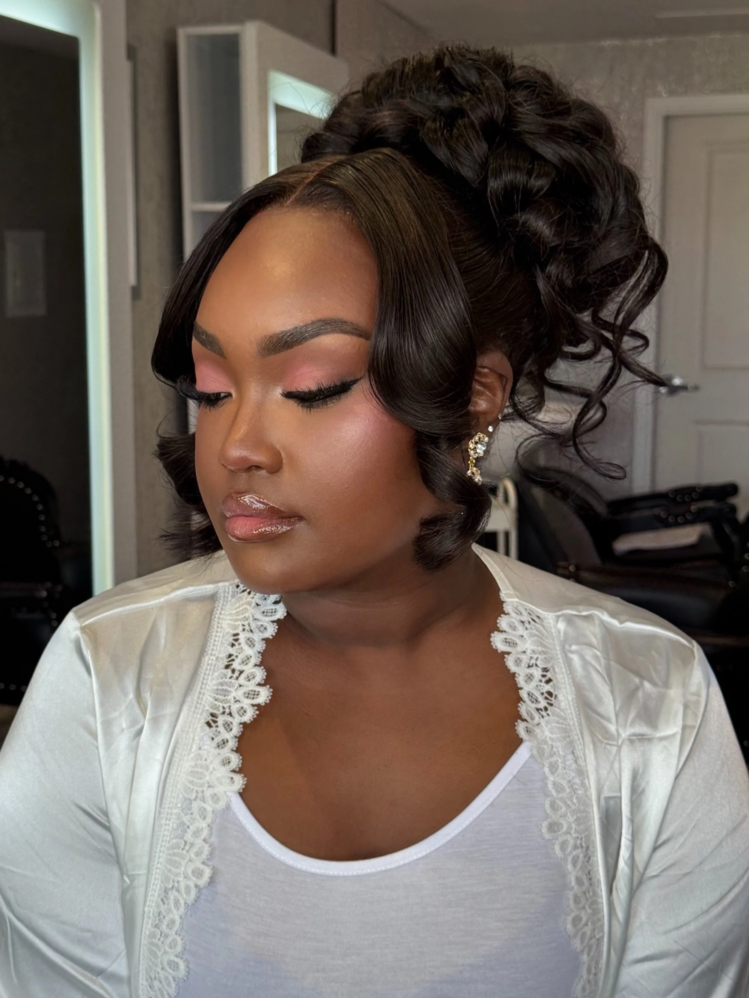 A woman with styled hair and makeup wearing a white top with lace detailing, and decorative earrings, standing in a room with a mirror and a door visible in the background.