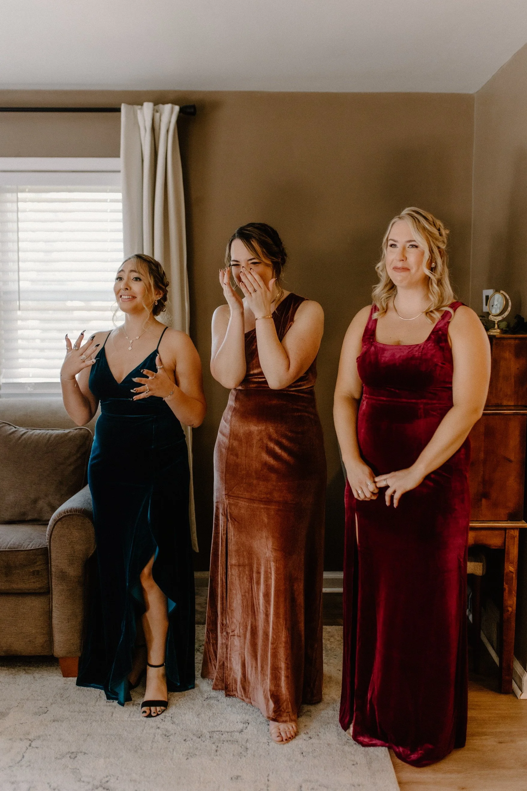 Bridesmaids in formal dresses standing together in a living room, smiling and laughing before first look.