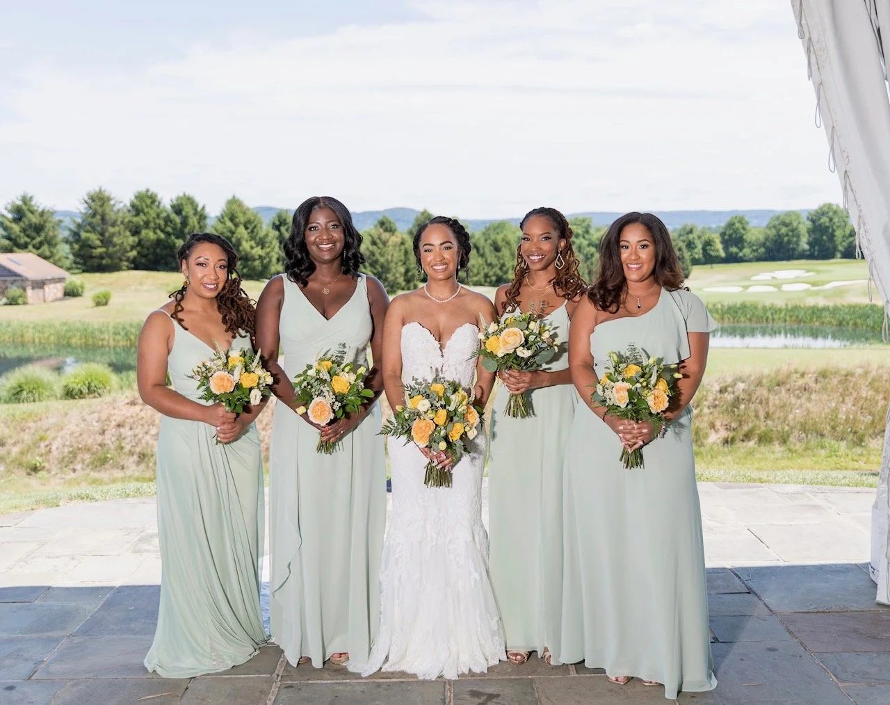 Bride and bridesmaids at a wedding outdoors, holding bouquets of yellow and white flowers, with a lush green landscape and golf course in the background.