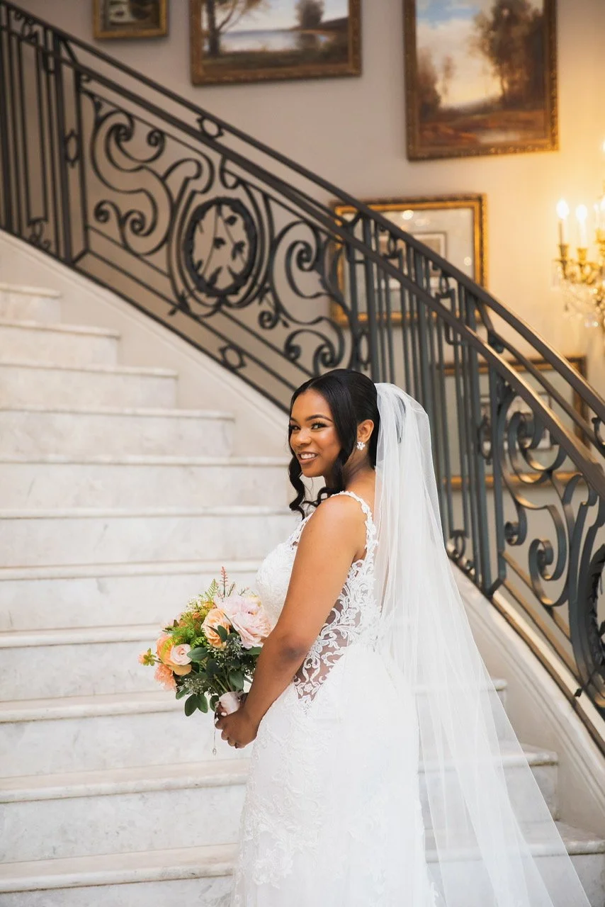 Bride in wedding dress holding bouquet standing on marble staircase with black wrought iron railing in elegant indoor setting.