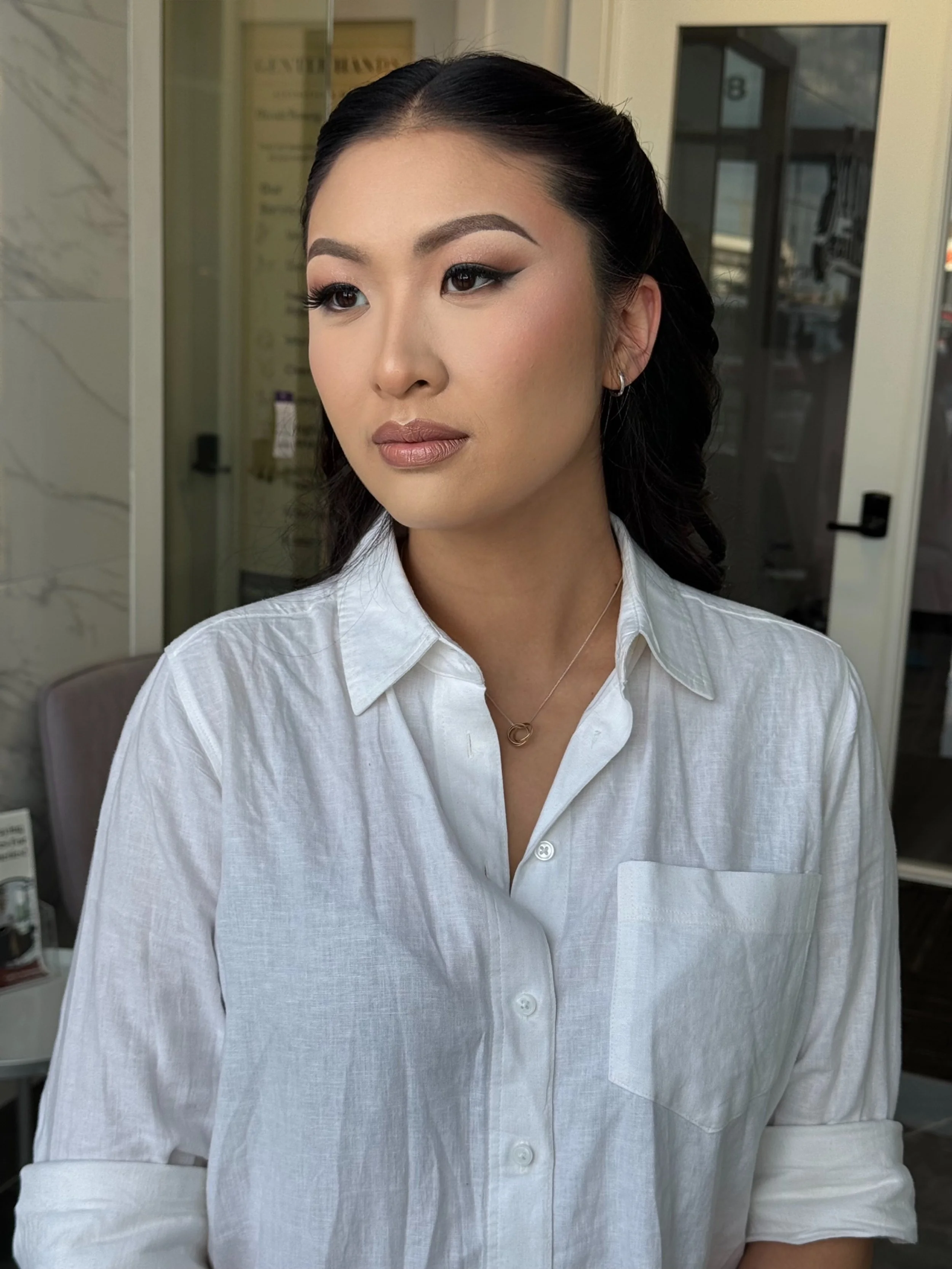 A woman with dark hair styled in waves, wearing makeup including eyeliner and lipstick, dressed in a white button-up shirt, standing indoors near a glass door with a marble wall behind her.