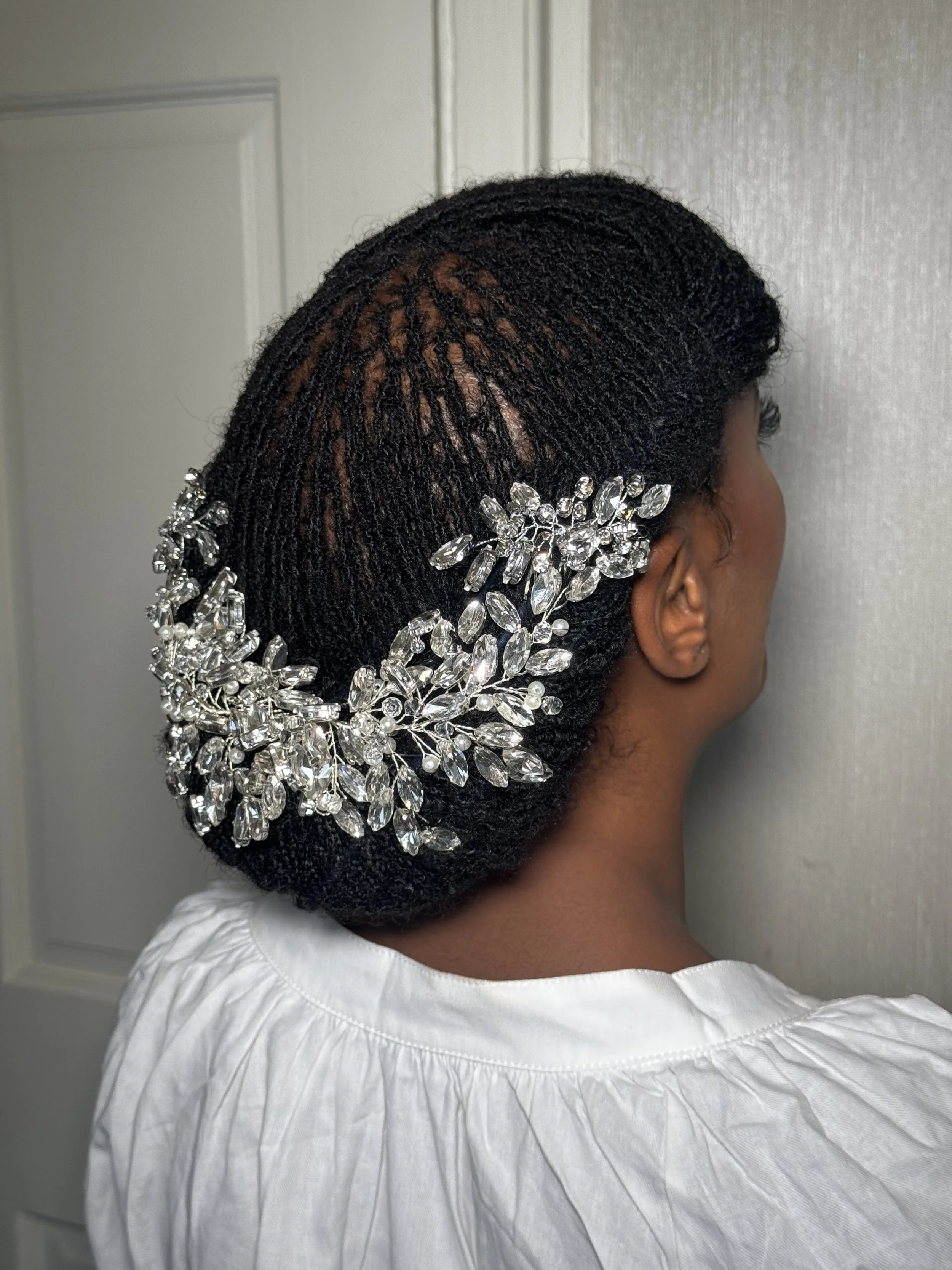 Close-up of a woman's head showcasing intricate black textured hair styled with a silver floral hair accessory and a white top with gathered fabric.