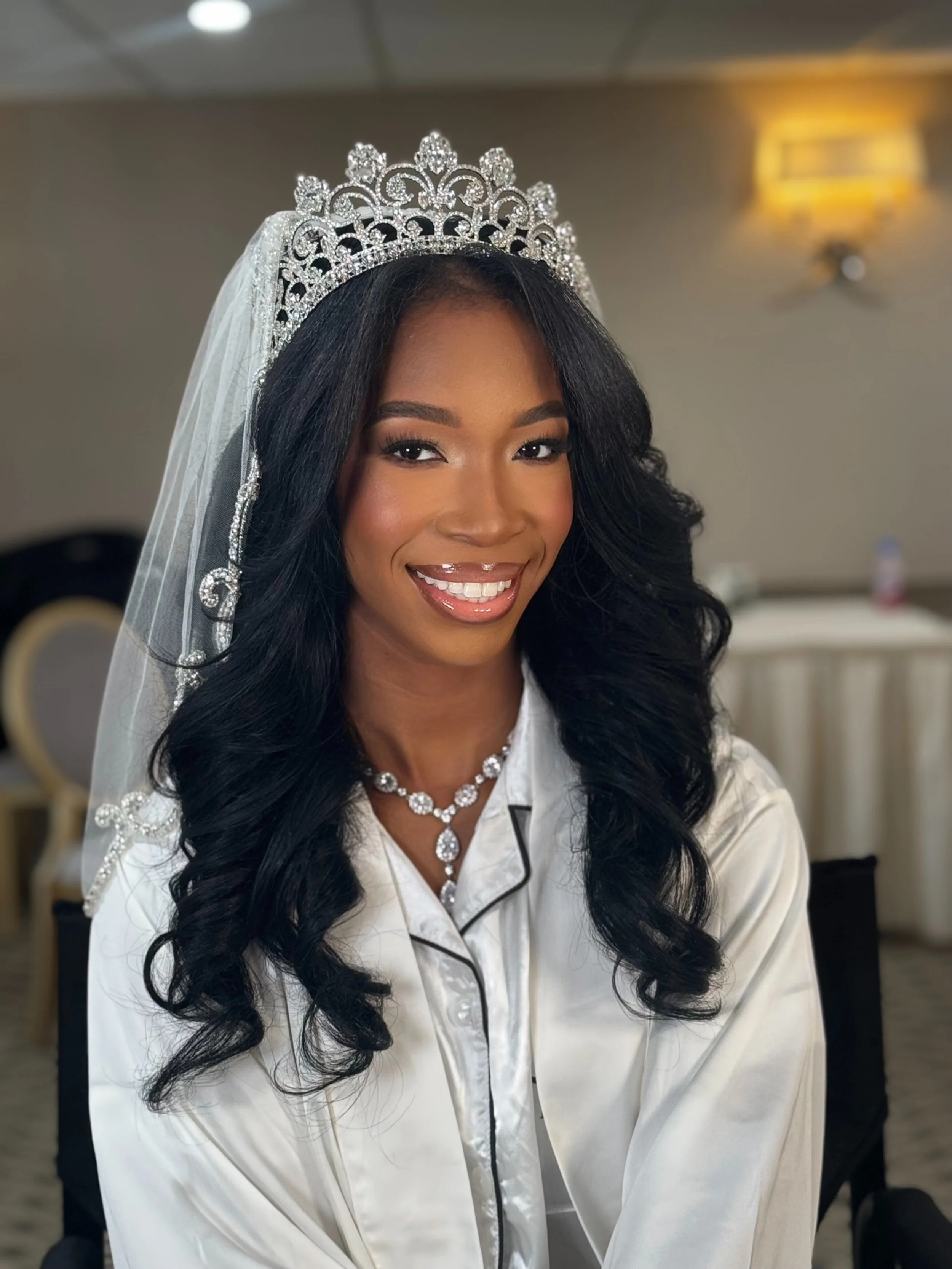 A woman with long black wavy hair is wearing a silver tiara with rhinestones, a sheer white veil, and a pearl necklace. She is smiling and wearing satin pajamas with black piping.