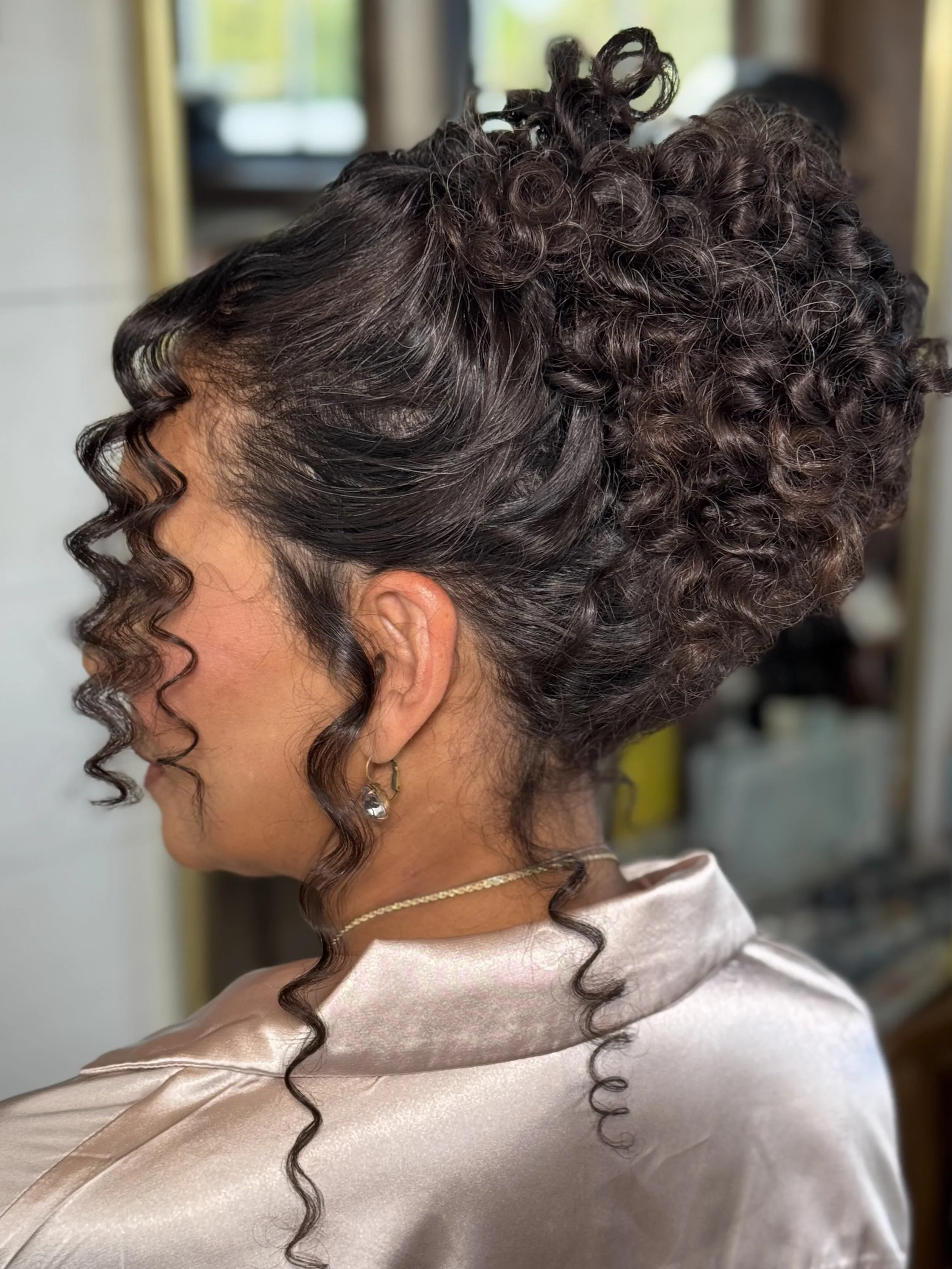 Side view of a woman with dark, curly hair styled in an elegant updo with loose curls framing her face, wearing a satin-like beige or champagne-colored top and jewelry.