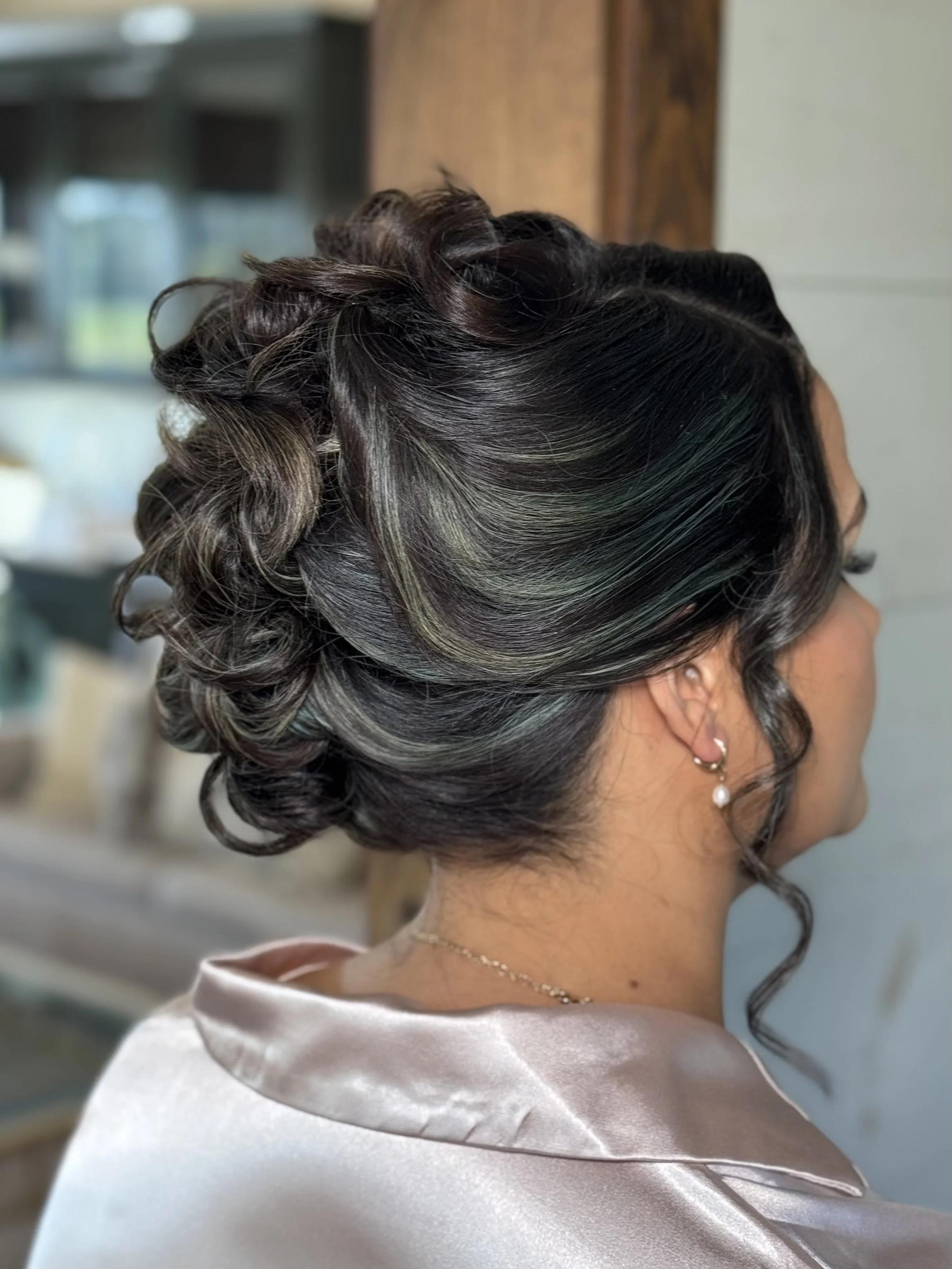 A woman with a short, styled, curls updo hairstyle with dark hair and subtle highlights, wearing pearl earrings and a satin clothing.