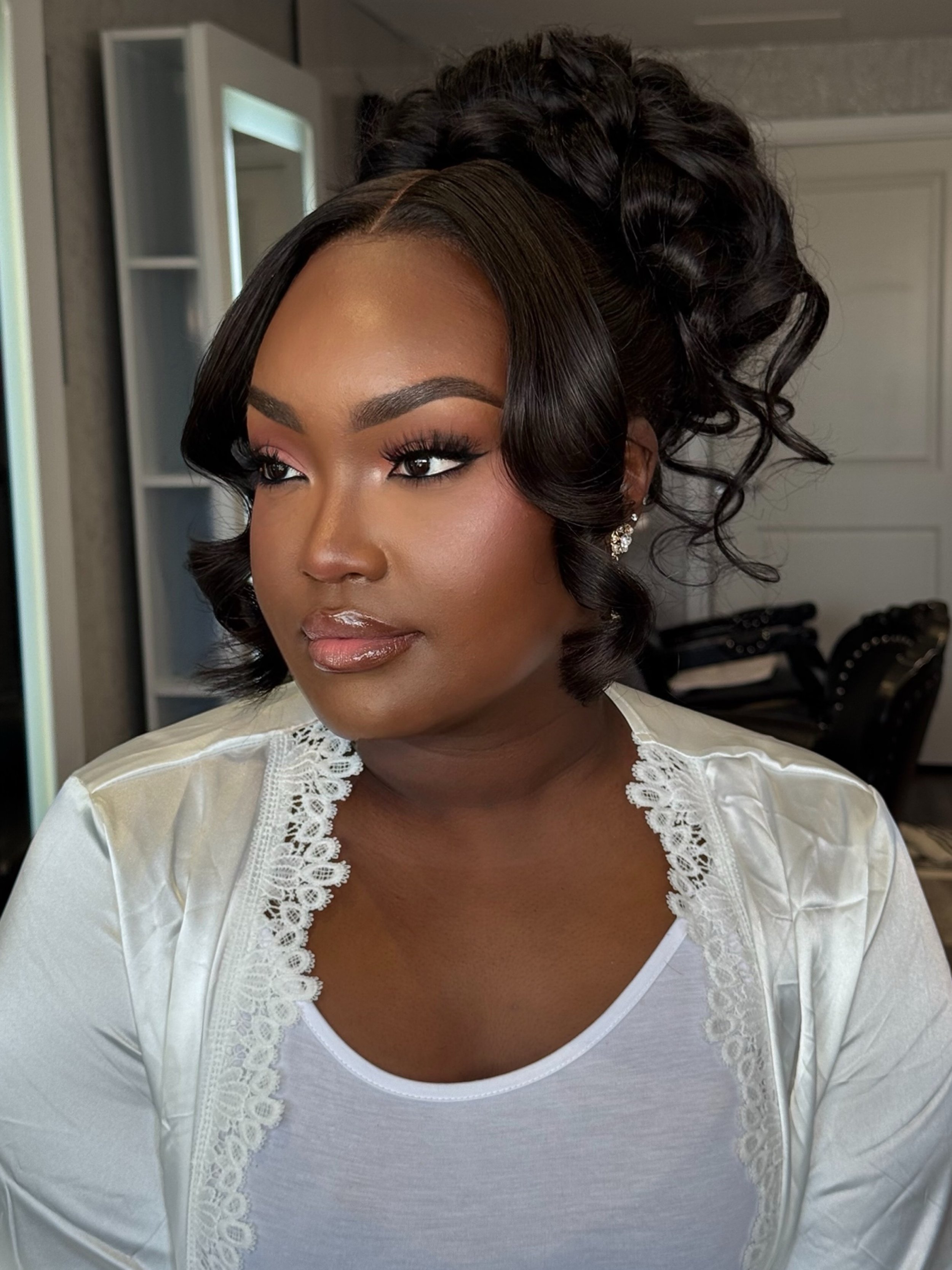 Close-up of a woman with expertly styled dark, curly hair, wearing a white top with lace details, and earrings, in a room with a mirror and clothing in the background.