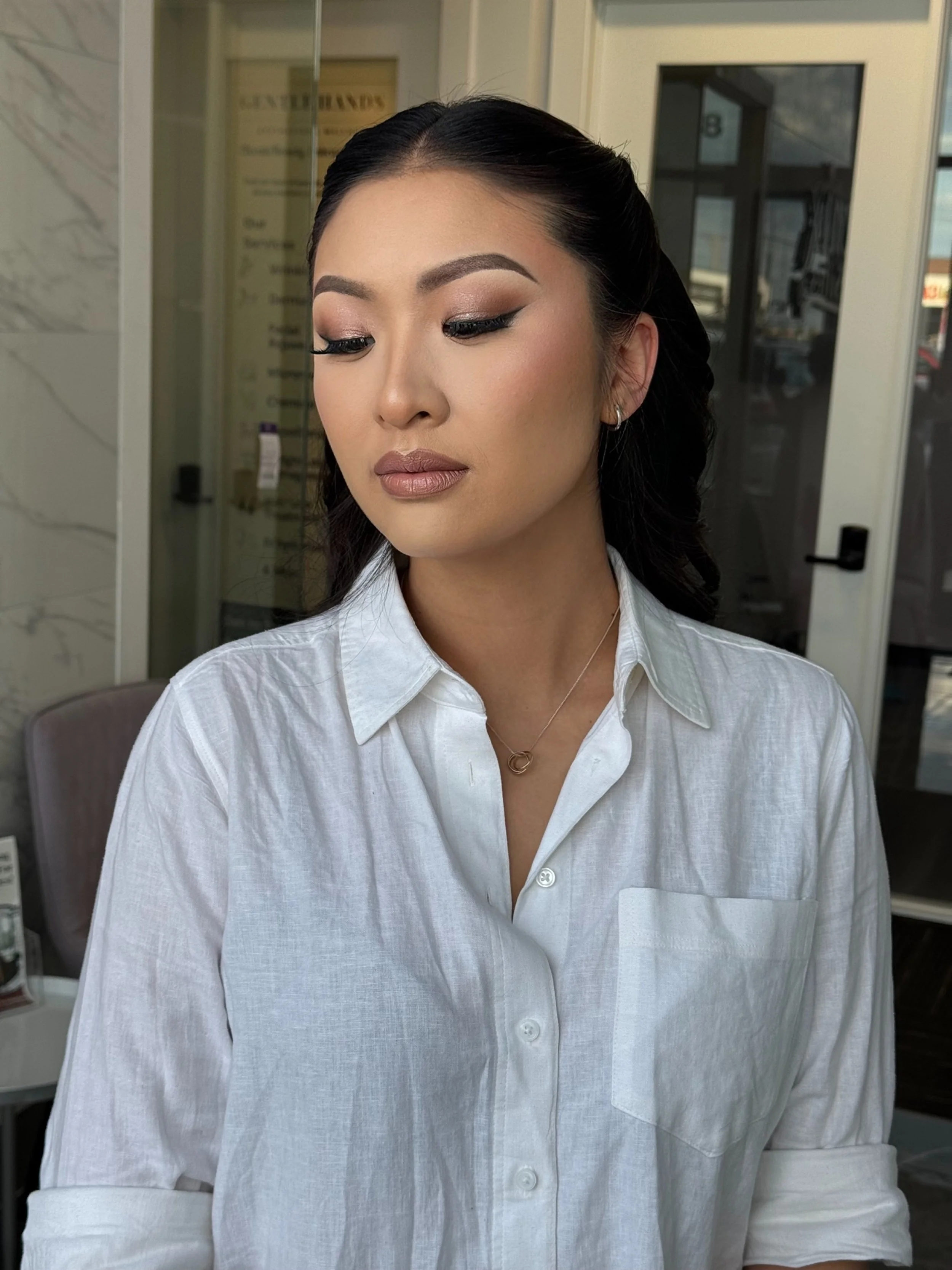 Close-up of a woman with dark hair styled in waves, wearing makeup with defined eyebrows, eyeliner, and nude lipstick, dressed in a white button-up shirt, standing indoors near a glass door.